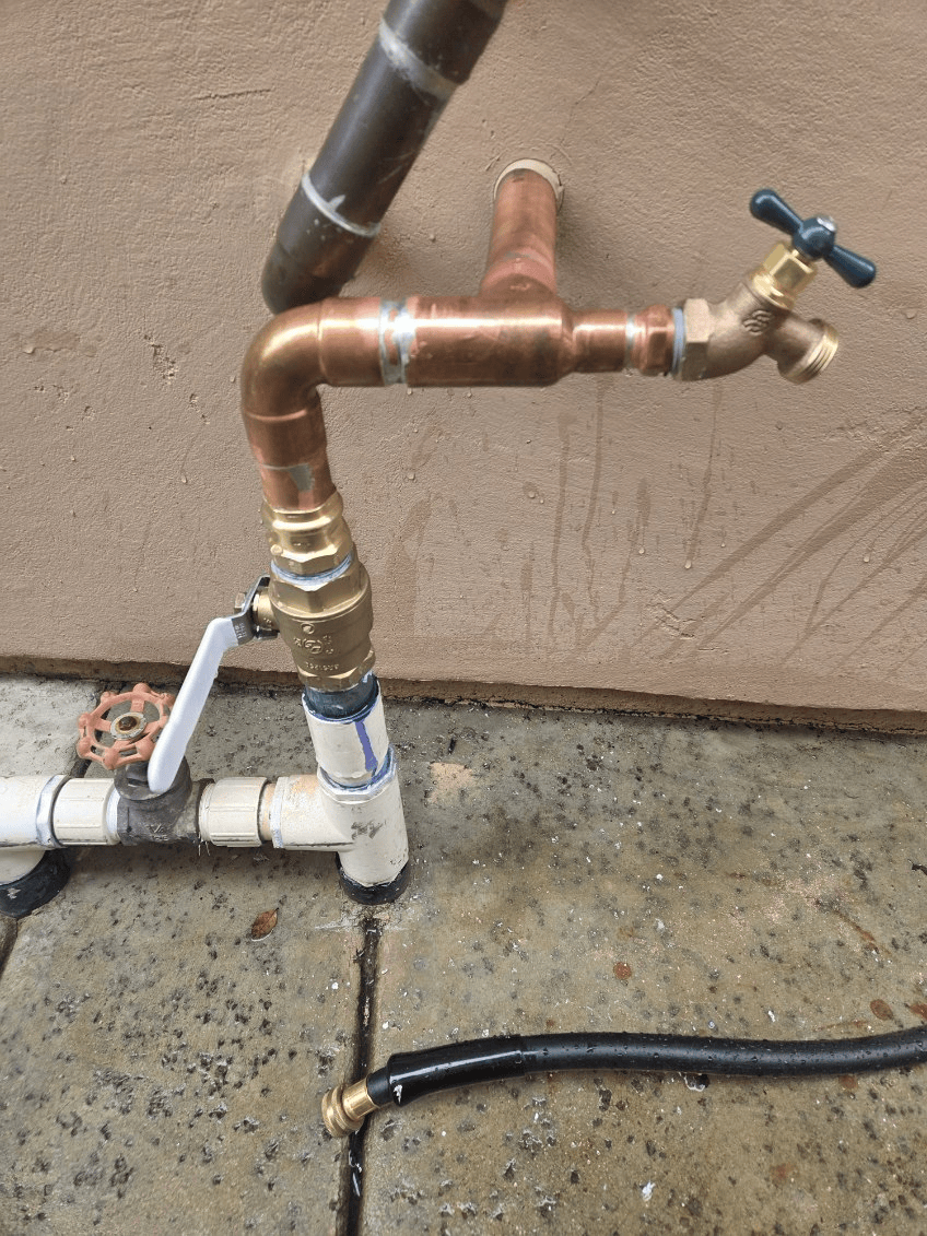 Outdoor water faucet with copper and PVC pipes connected to a wall, black hose on the ground.