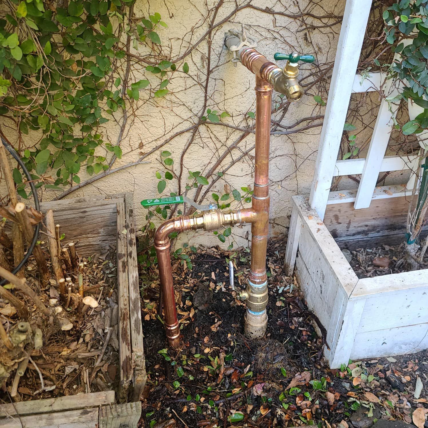 Copper water pipes with two faucets in a garden setting, against a light-colored wall with climbing vines.