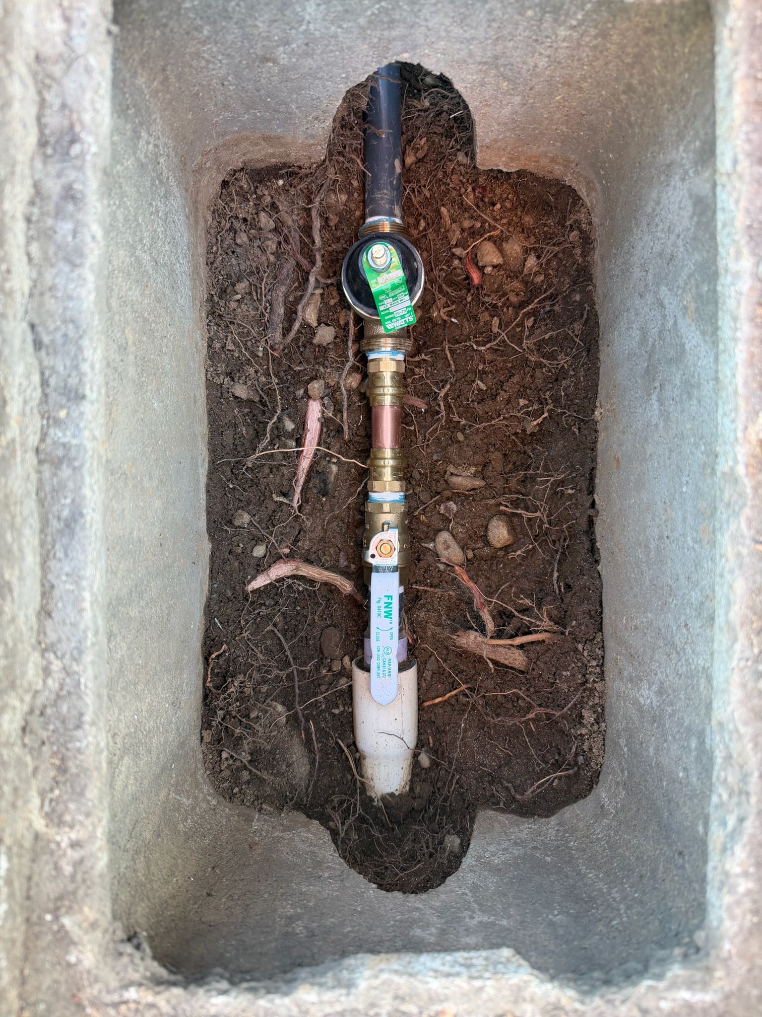 Water meter with exposed pipe inside a concrete utility box, surrounded by soil and roots.