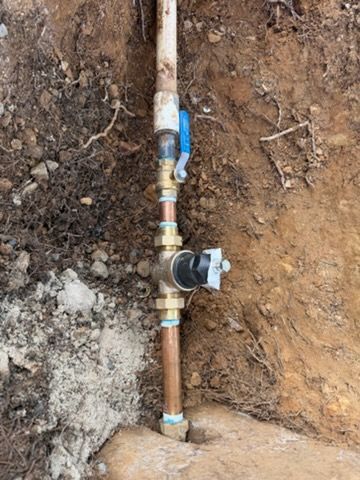 An outdoor water pipe installation in a dirt trench featuring a blue shut-off valve and a pressure regulator.