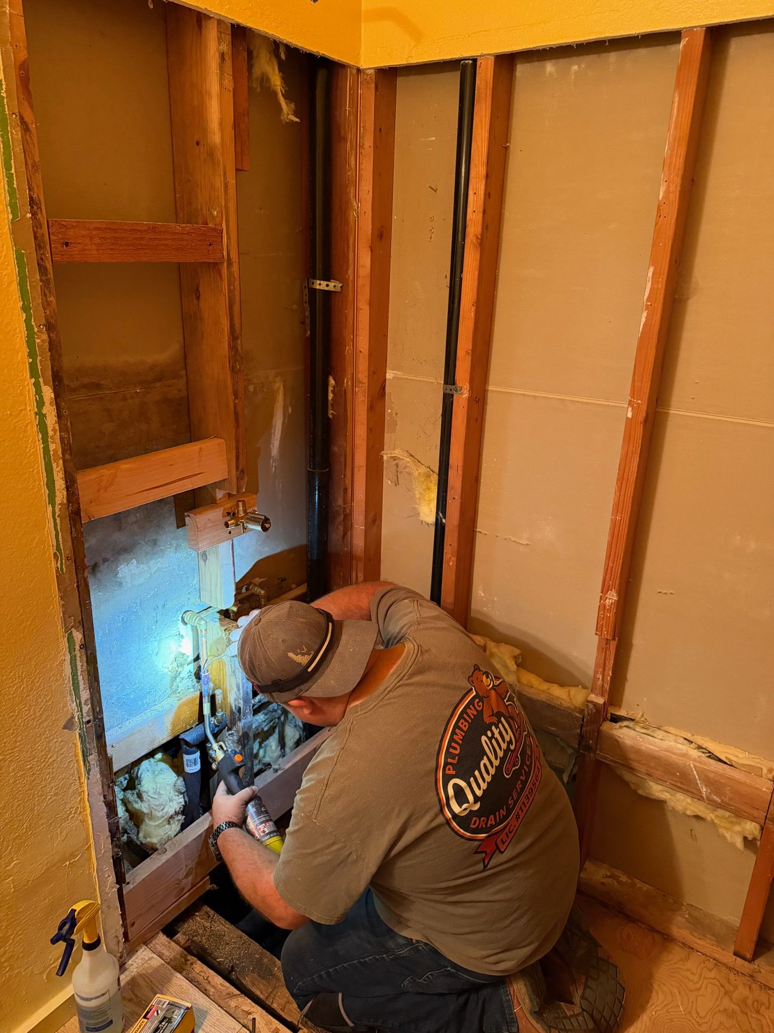 Worker kneeling in a bathroom wall opening, repairing plumbing with a flashlight in a stripped room