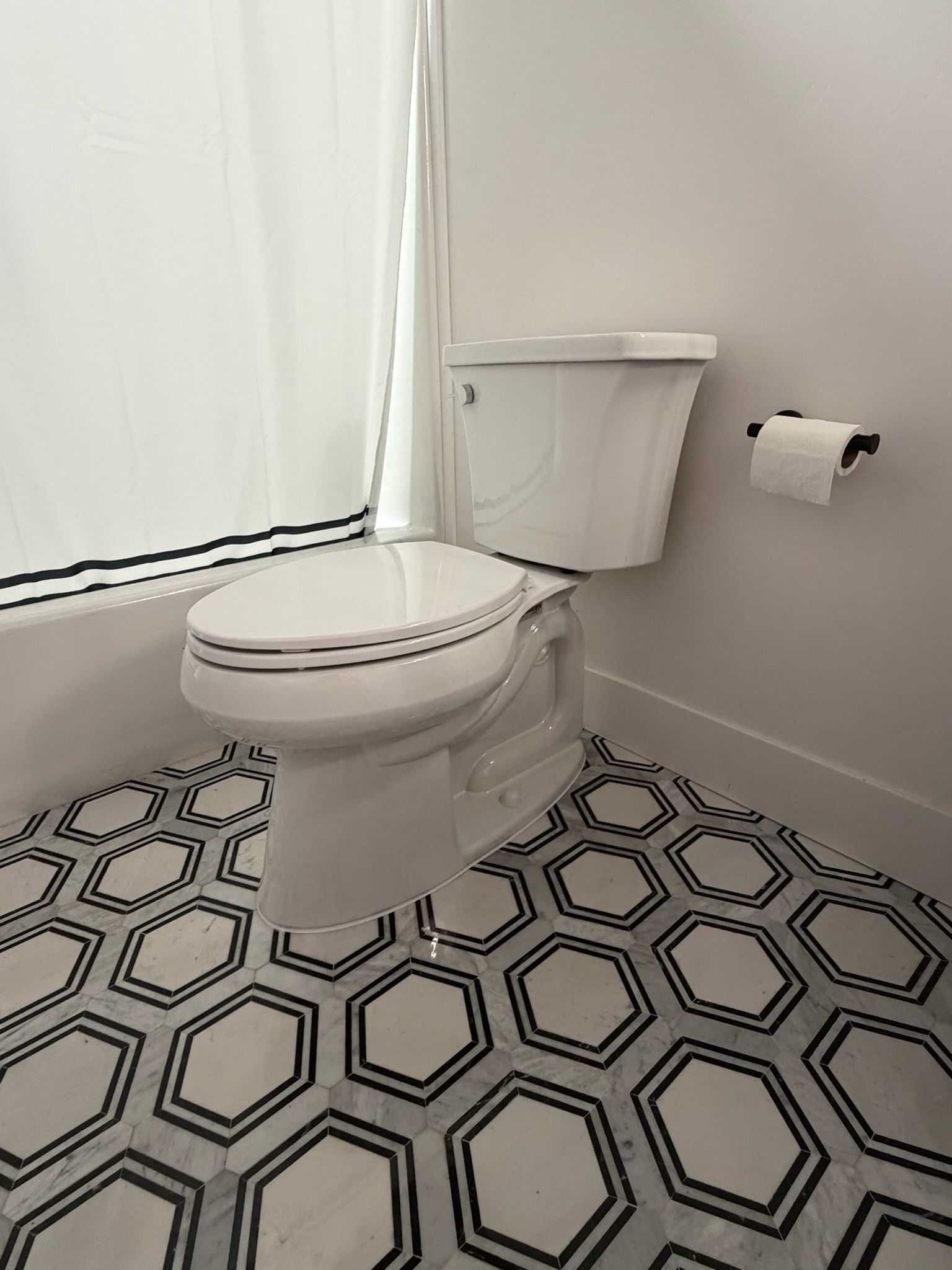 Small bathroom with a toilet, patterned black-and-white floor, white walls, and a window with a curtain.