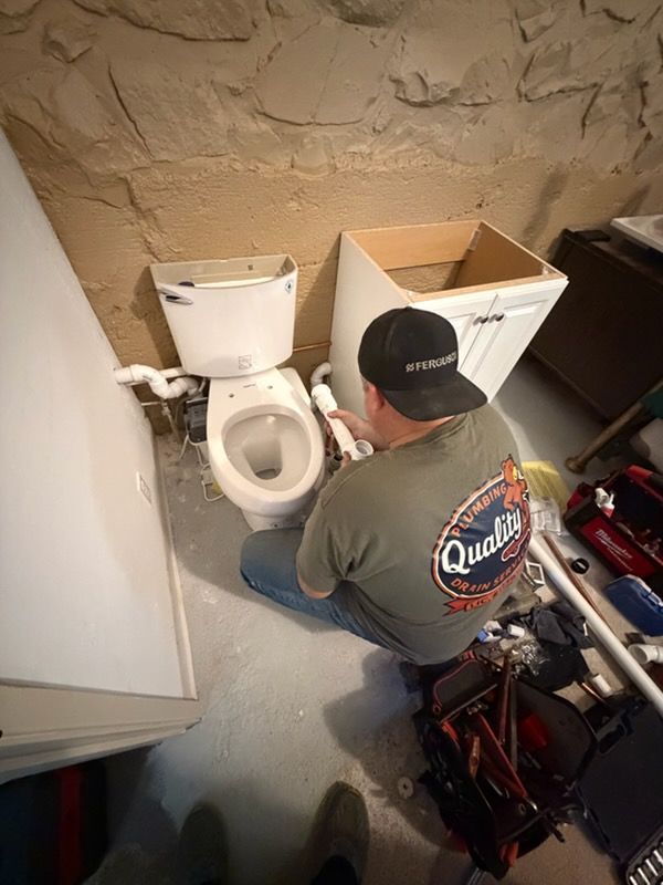A plumber kneels on a concrete floor to install white PVC plumbing pipes connected to a toilet in a basement bathroom.