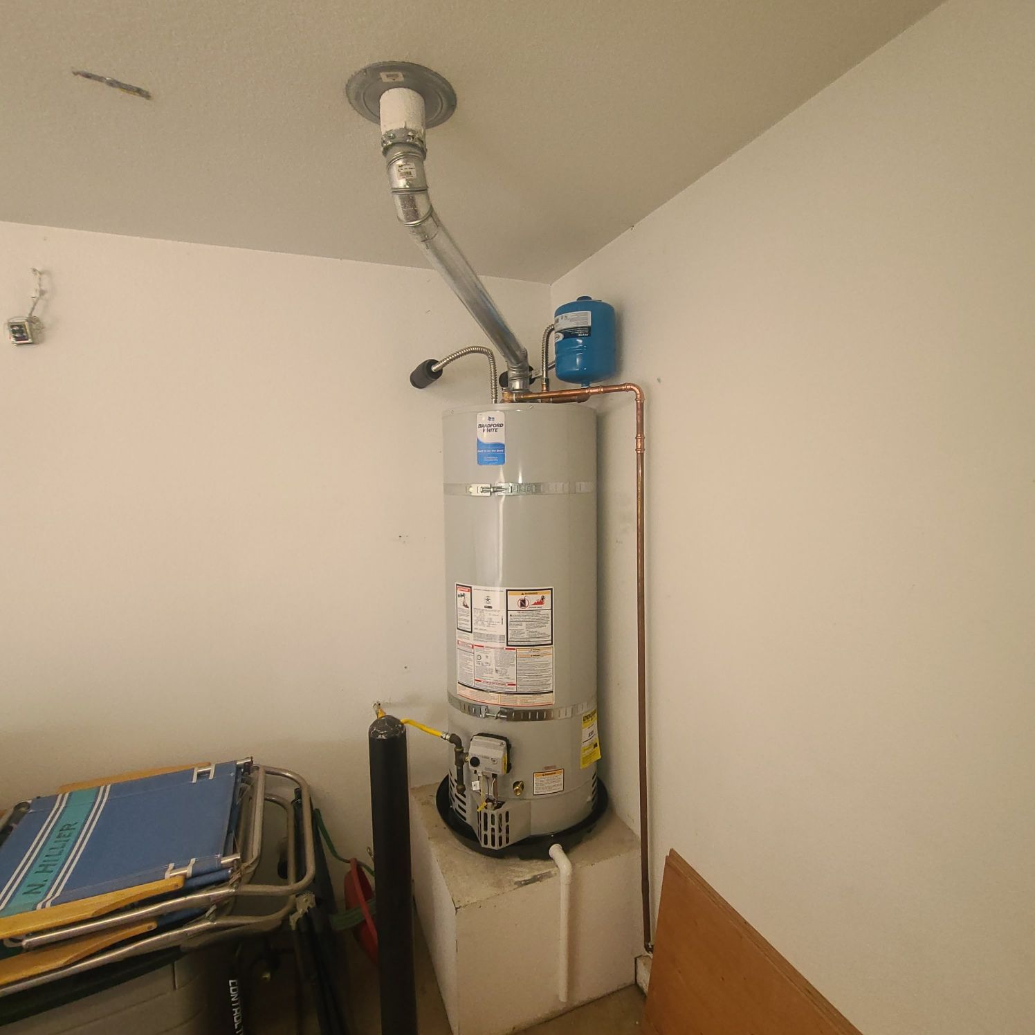 A water heater with a vent pipe in a corner of a room. It has copper pipes and a blue expansion tank.