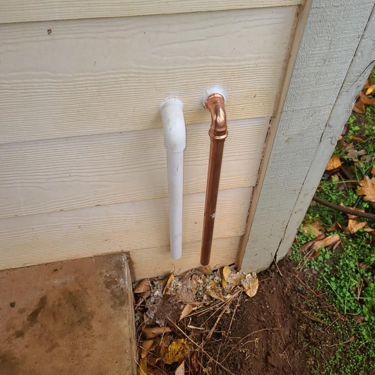 Two pipes protruding from the side of a building: copper and white, with the ground and grass visible.