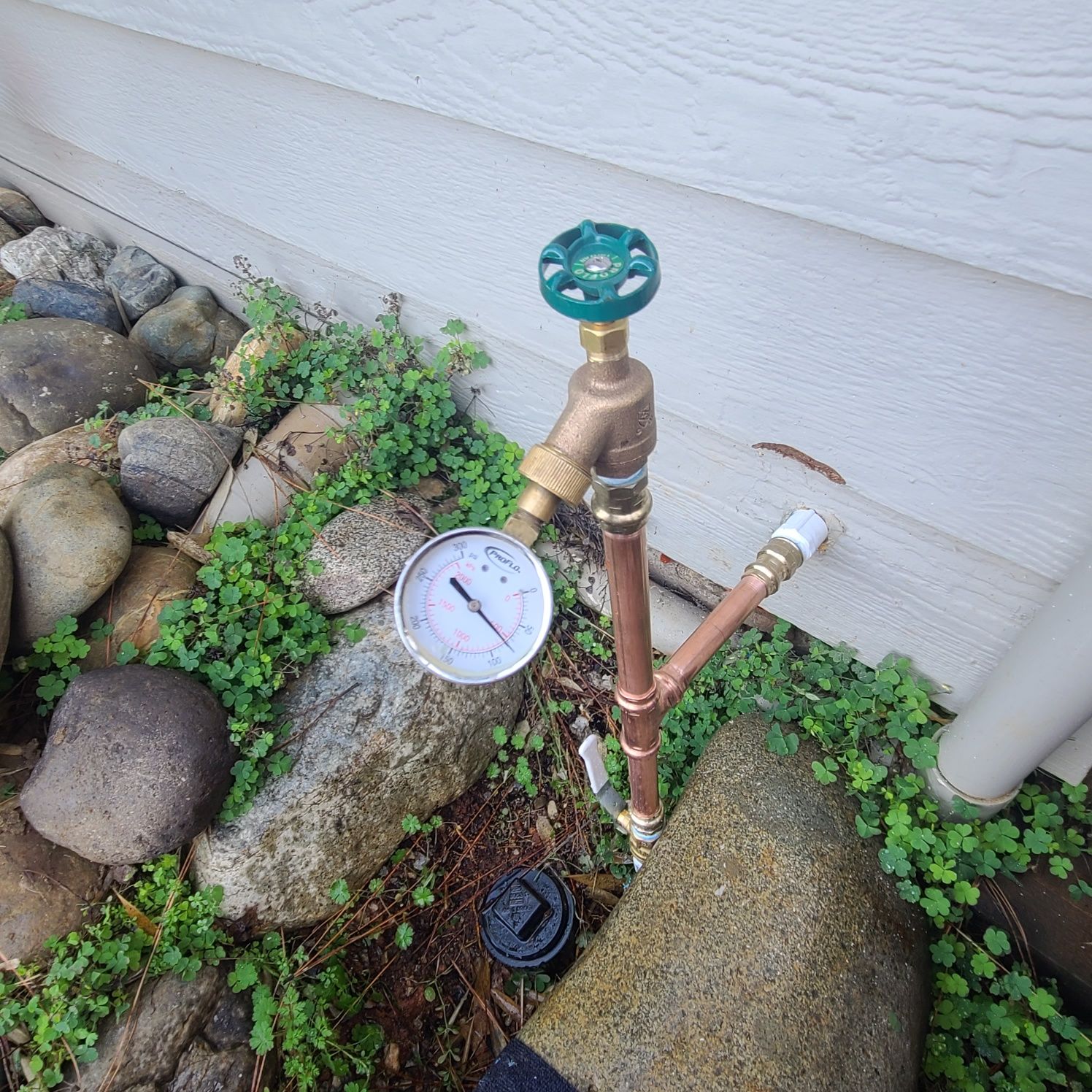Copper pipe with water pressure gauge and faucet near a house with rocks and weeds.