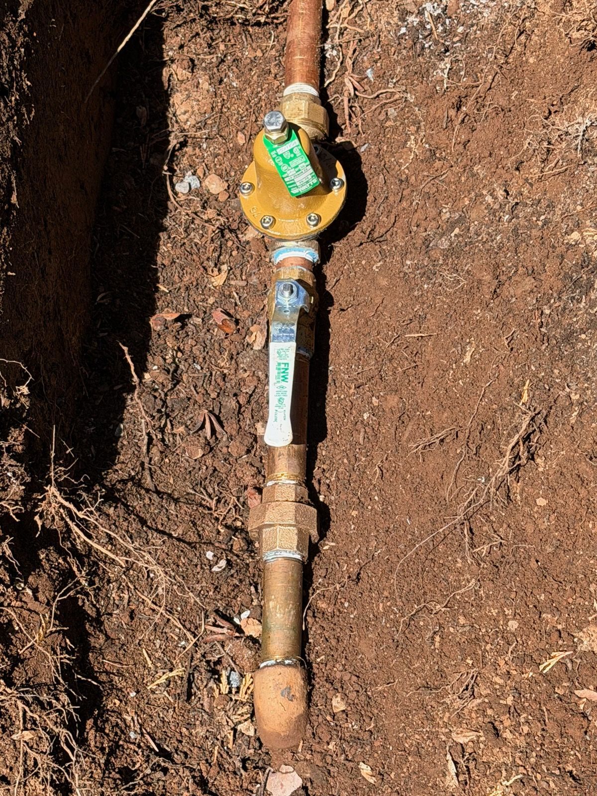 Gas pipe valve with yellow handle installed in a dirt trench and marked with green tape