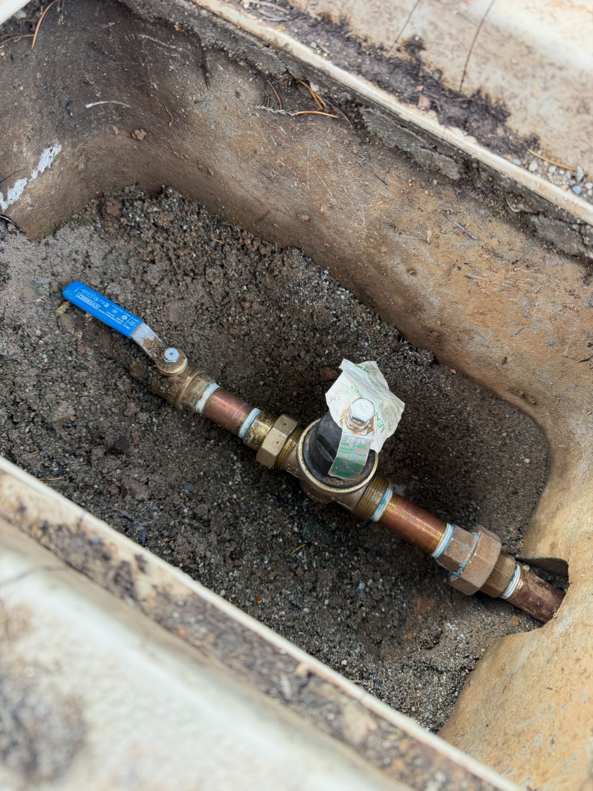 Brass water valve with blue handle, in a rectangular underground box, covered in dirt.