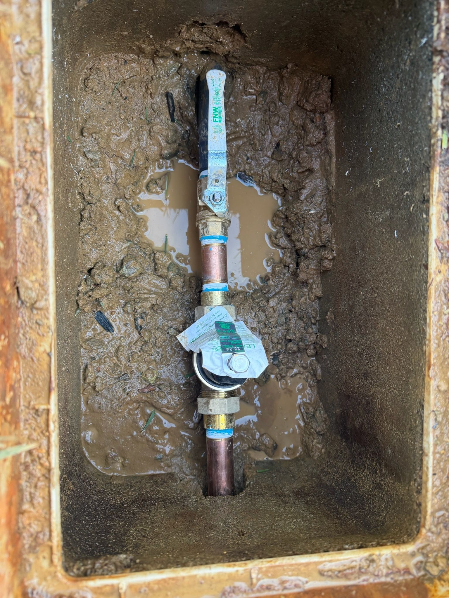 Water meter and valve in a muddy rectangular box. Copper pipes, blue and green accents.