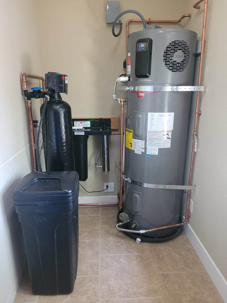 Water heater and water softener in a utility room. Gray tank, black filters, copper pipes, and a blue salt tank.