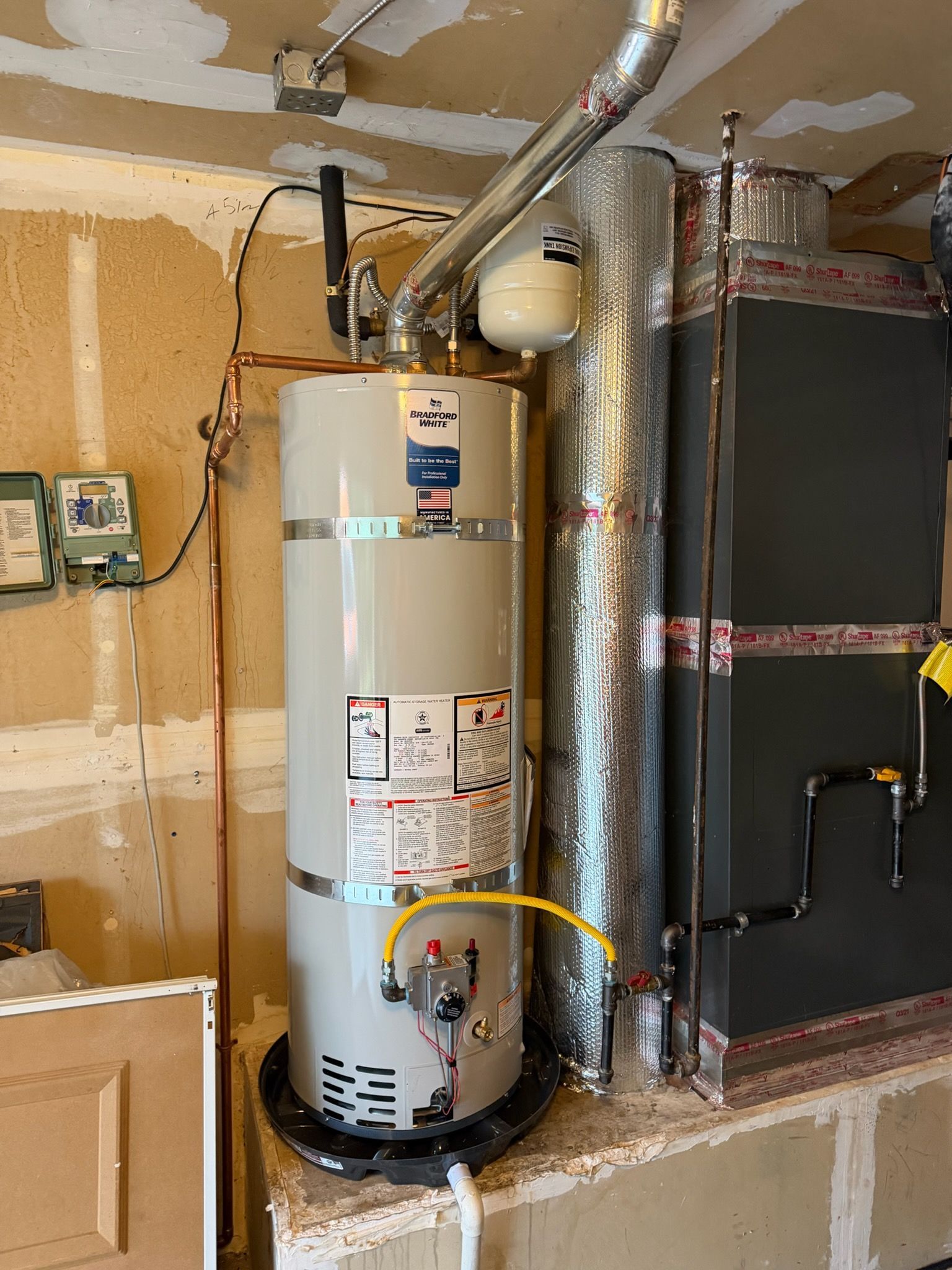 A tall, grey residential water heater sits in a drip pan on a raised platform next to a large metal HVAC unit.
