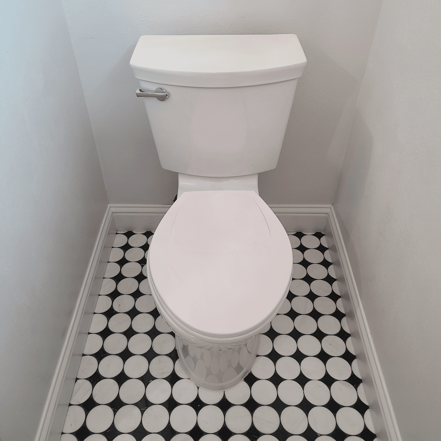 White toilet in a small bathroom with black and white circular tile flooring and white walls.