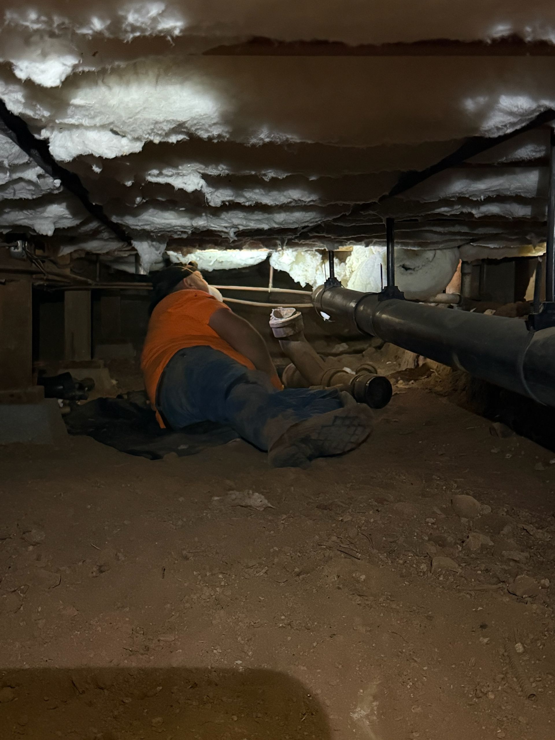 Worker in orange vest inspecting pipes in a low crawlspace with insulation overhead.
