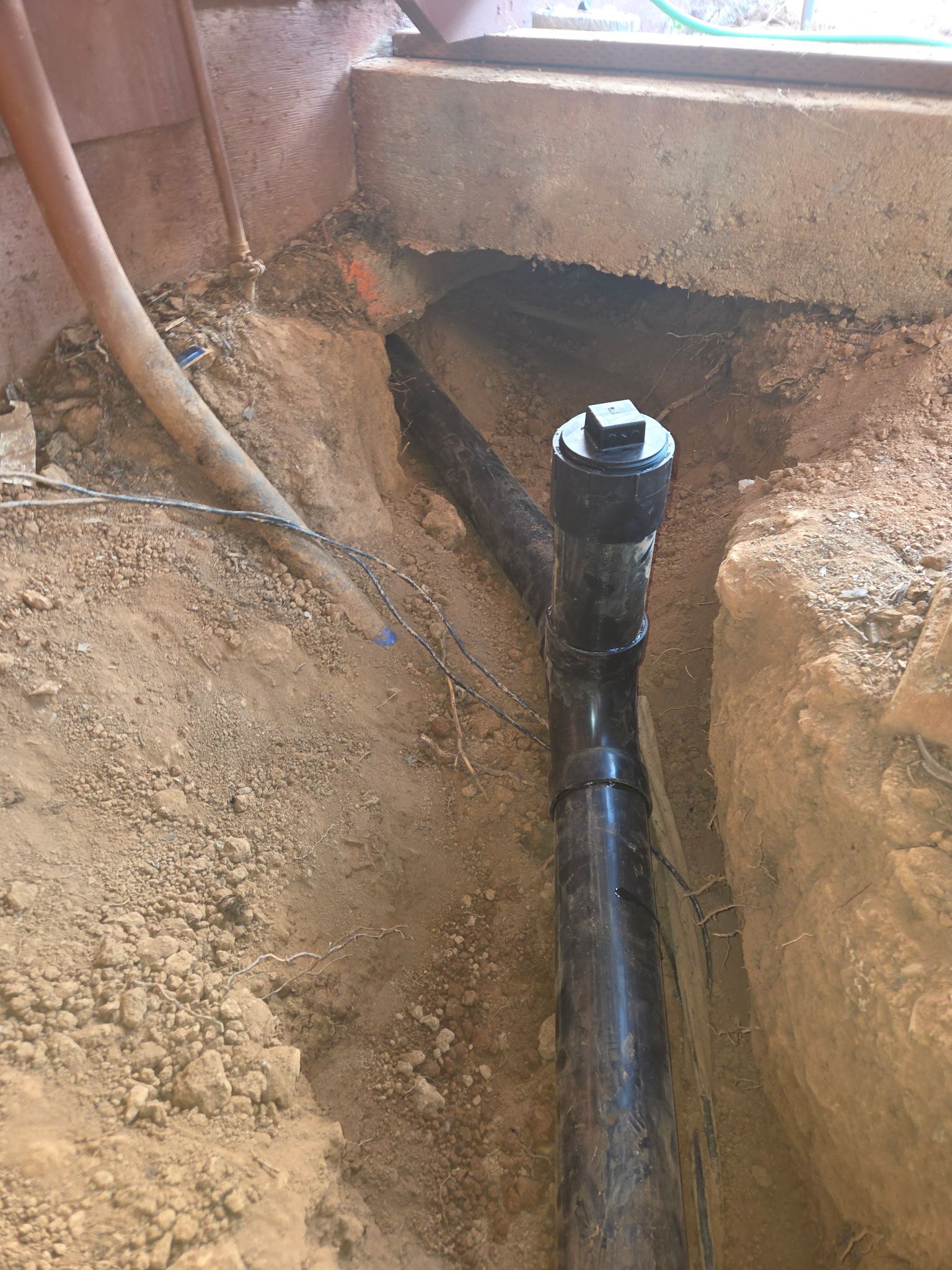 Black plumbing pipe in a dirt trench beside a concrete foundation drain area