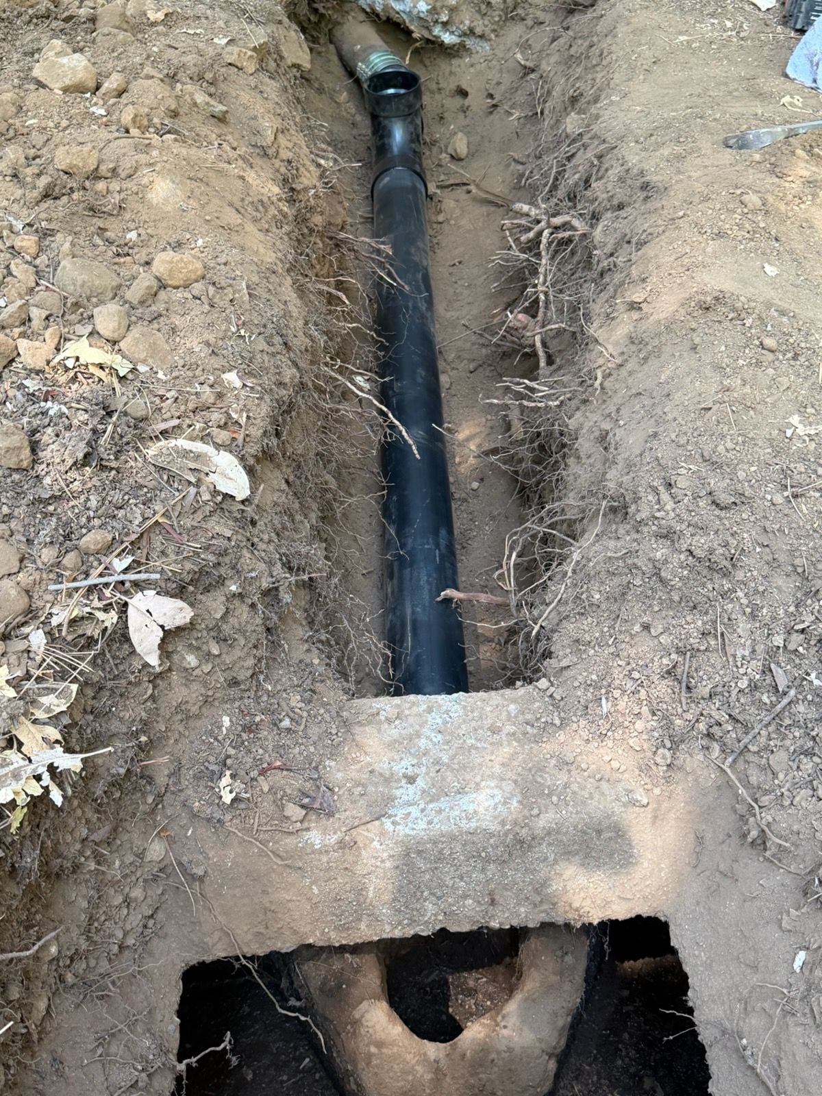 Trench with a black underground pipe running through dirt, crossing under a concrete slab