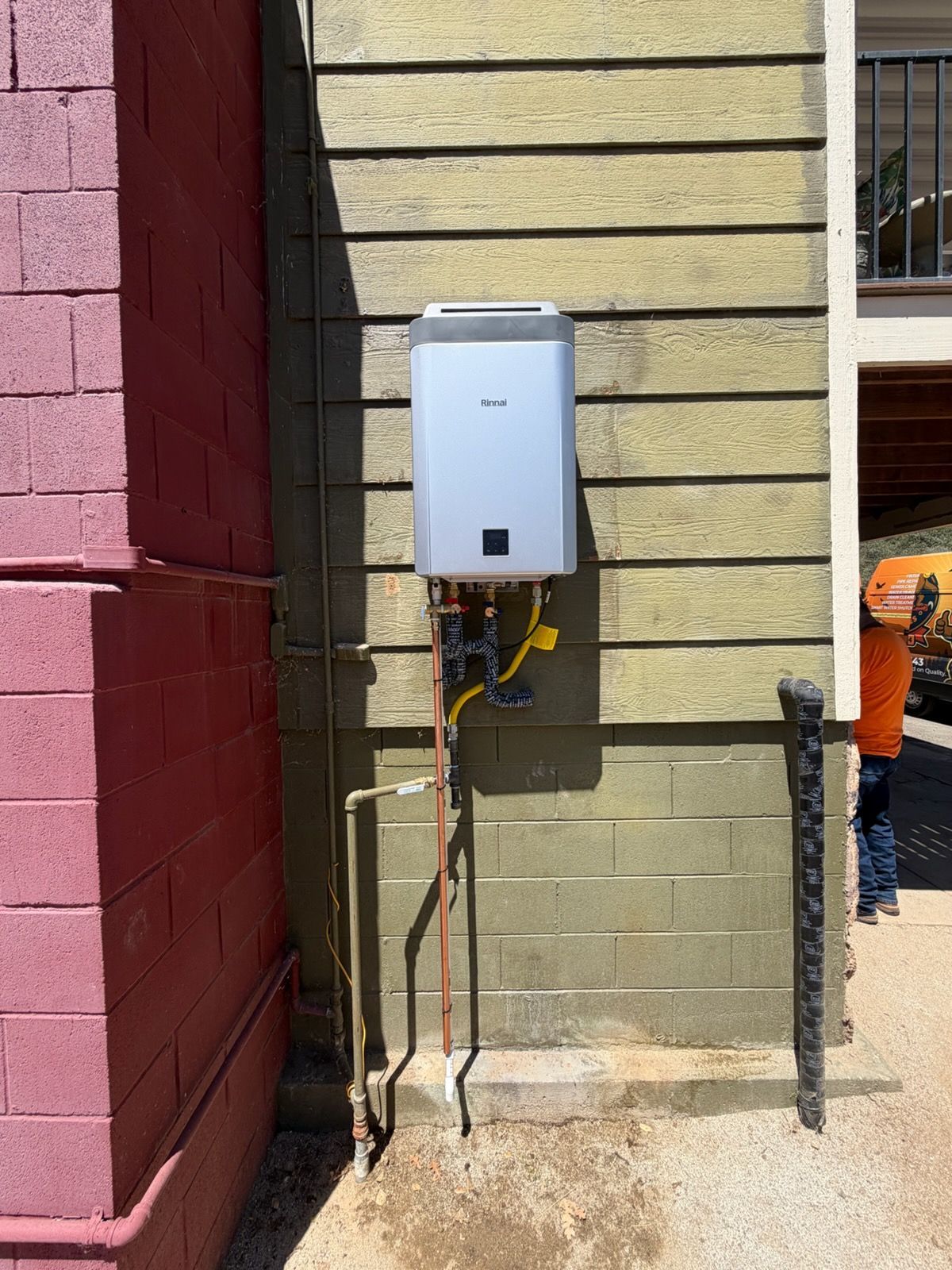 Wall-mounted utility box with exposed pipes on a beige exterior wall beside a red building corner.
