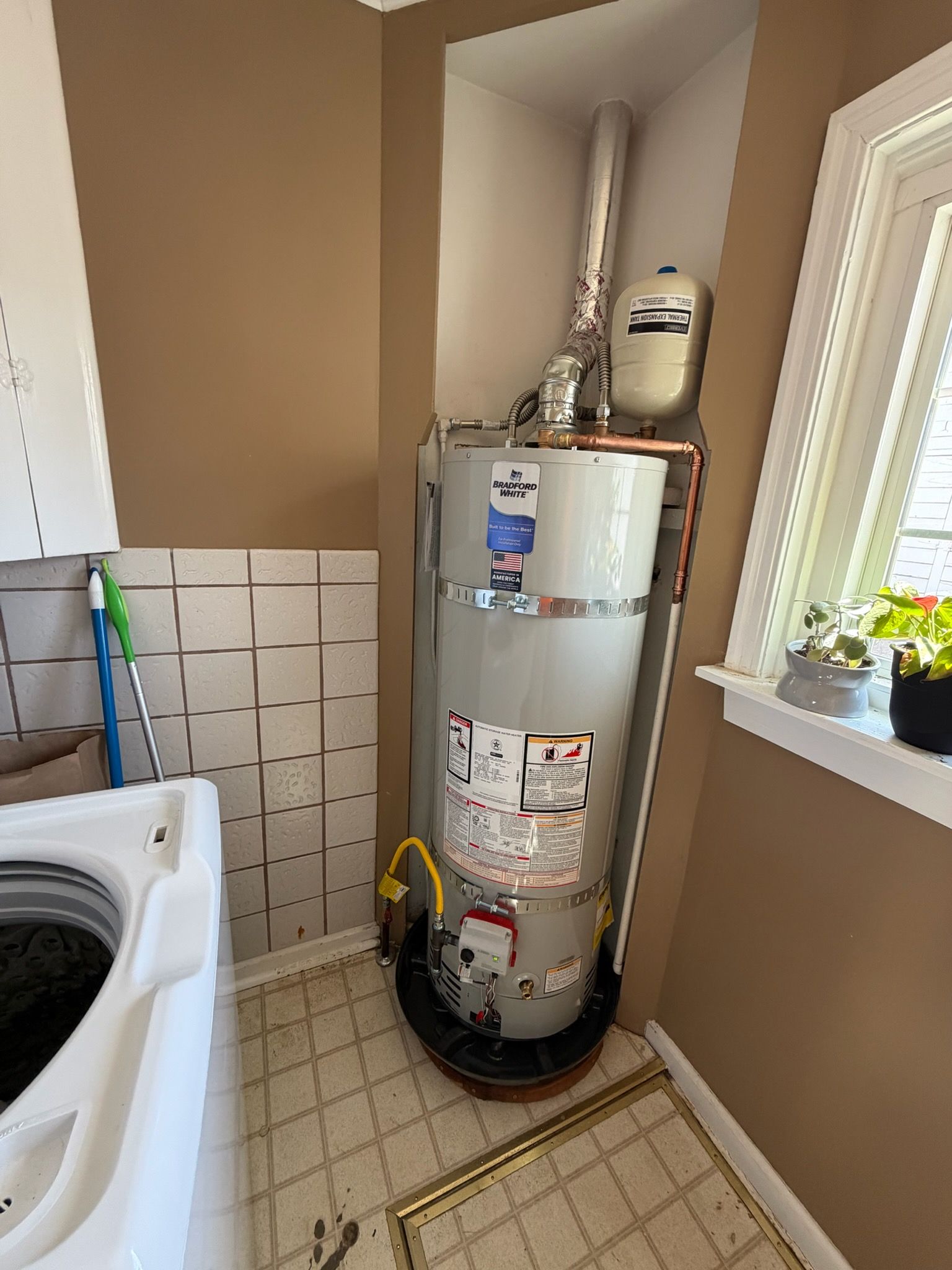 Water heater in a small laundry/utility nook beside a washing machine and window.