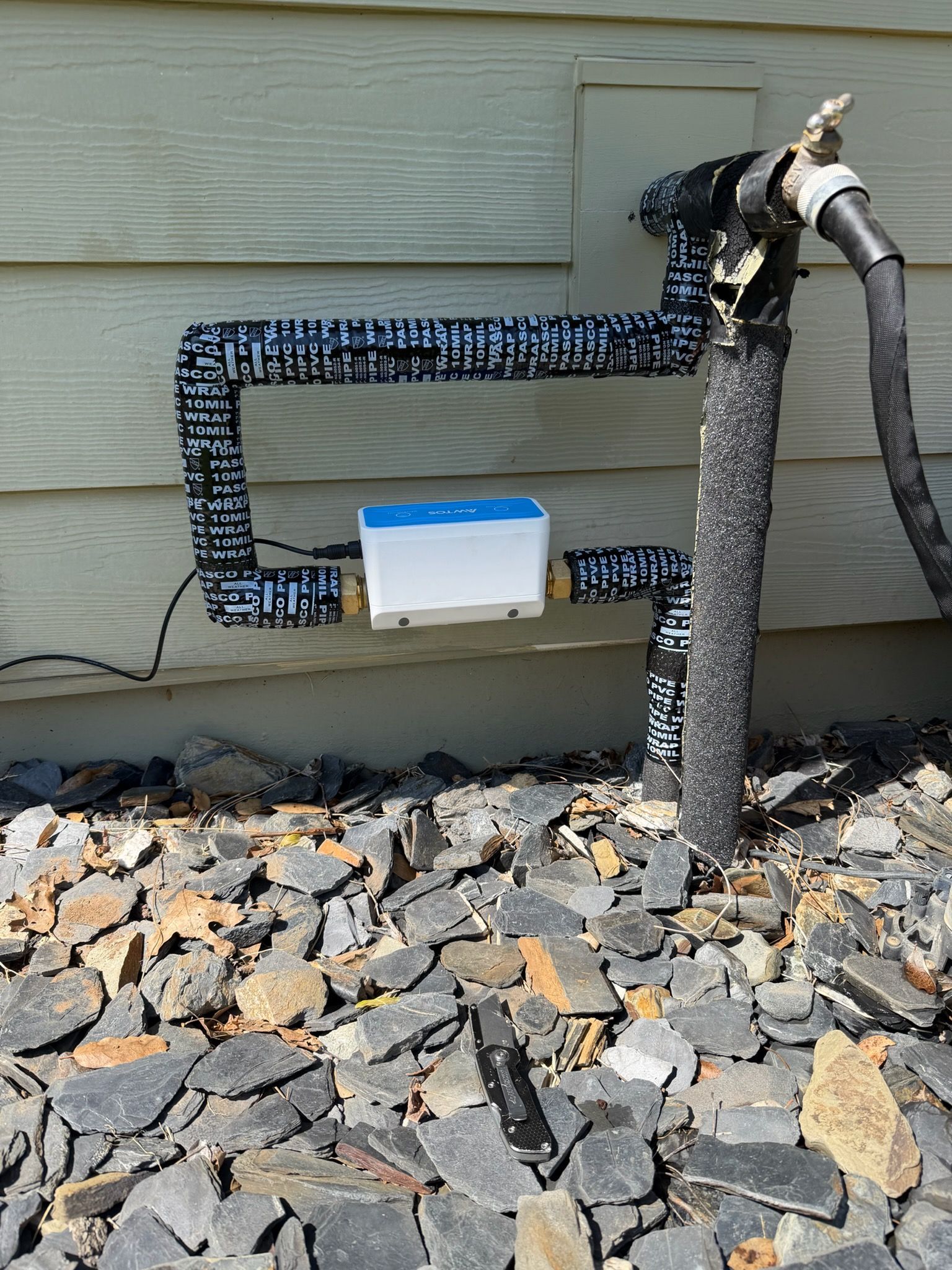 Black insulated outdoor pipes beside a house wall with a white utility box and hose on gravel