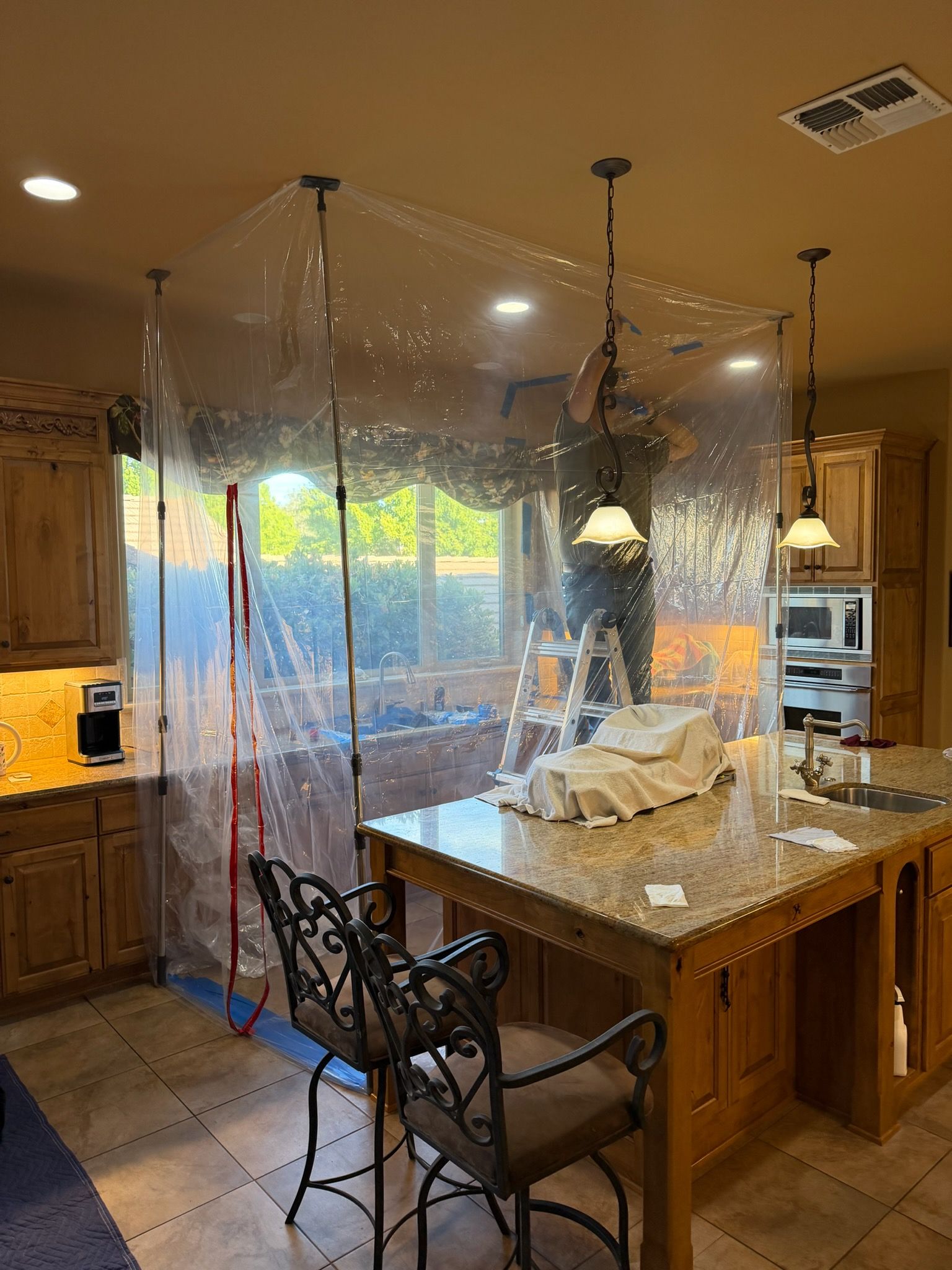 Kitchen island wrapped in clear plastic sheeting during renovation, with hanging lights and stools nearby