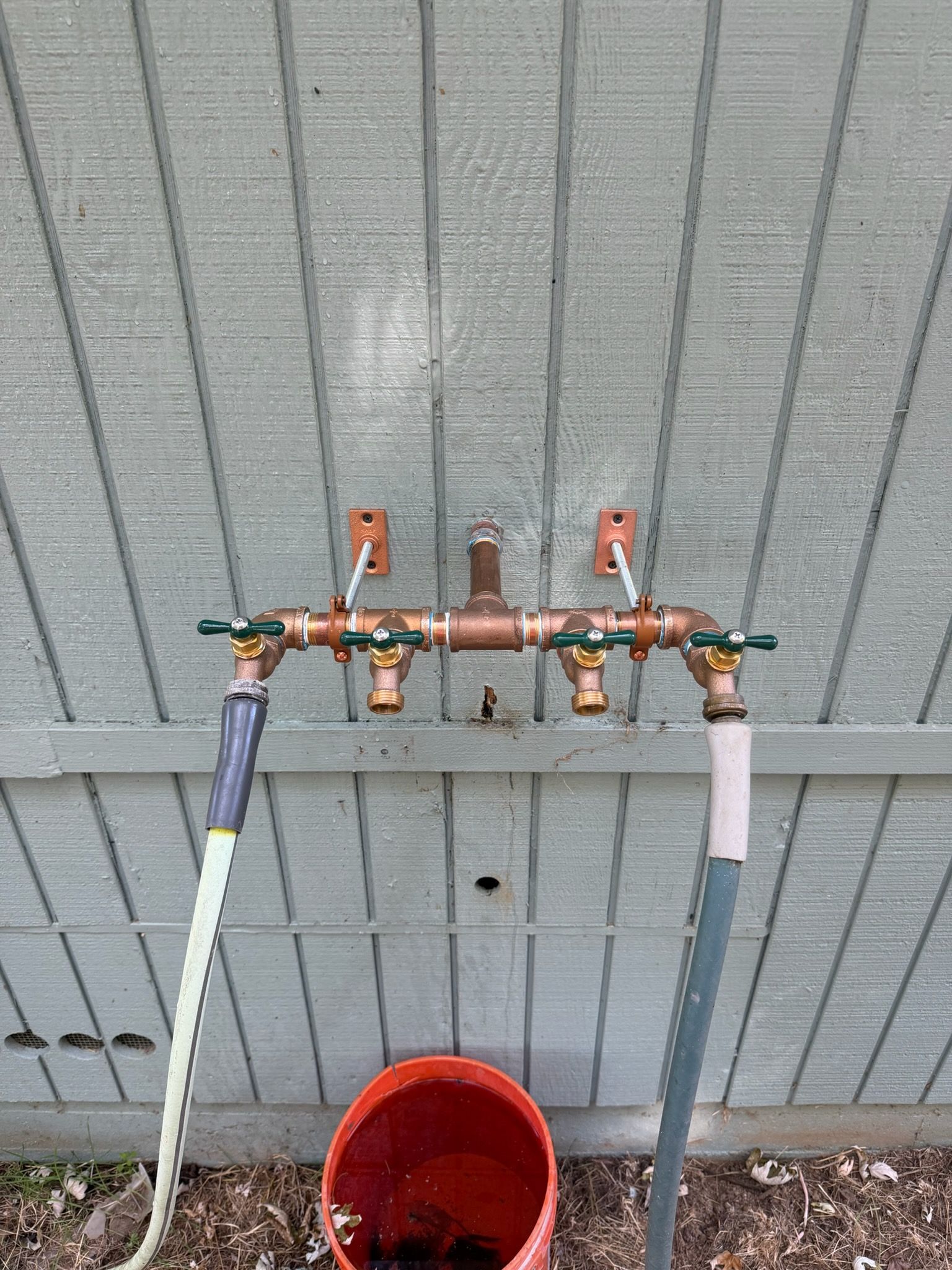 Outdoor utility pipe assembly with three shutoff valves and connected hoses against a gray wall, above a red bucket