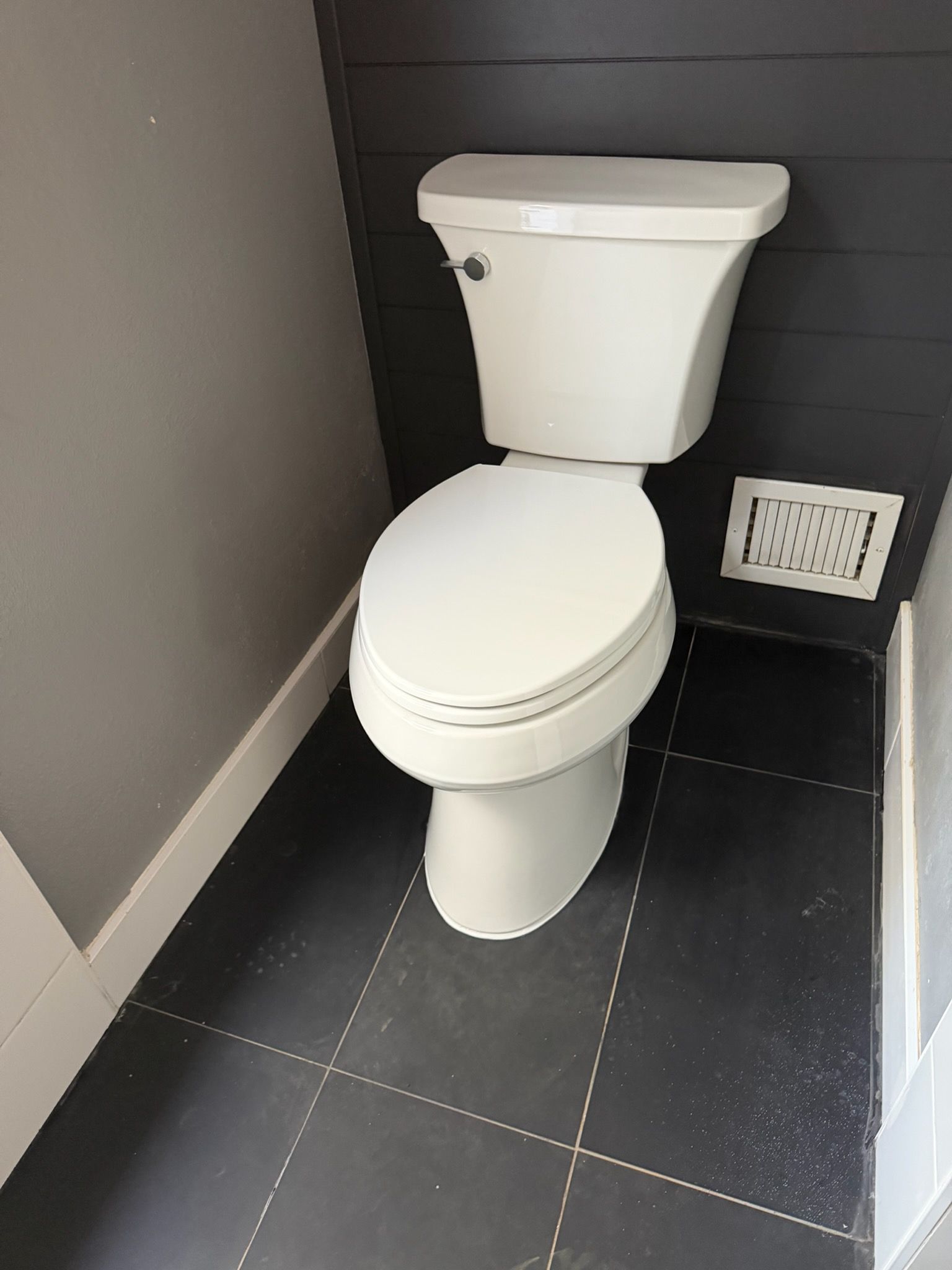 Small white toilet in a narrow restroom with dark tiled floor and gray walls.
