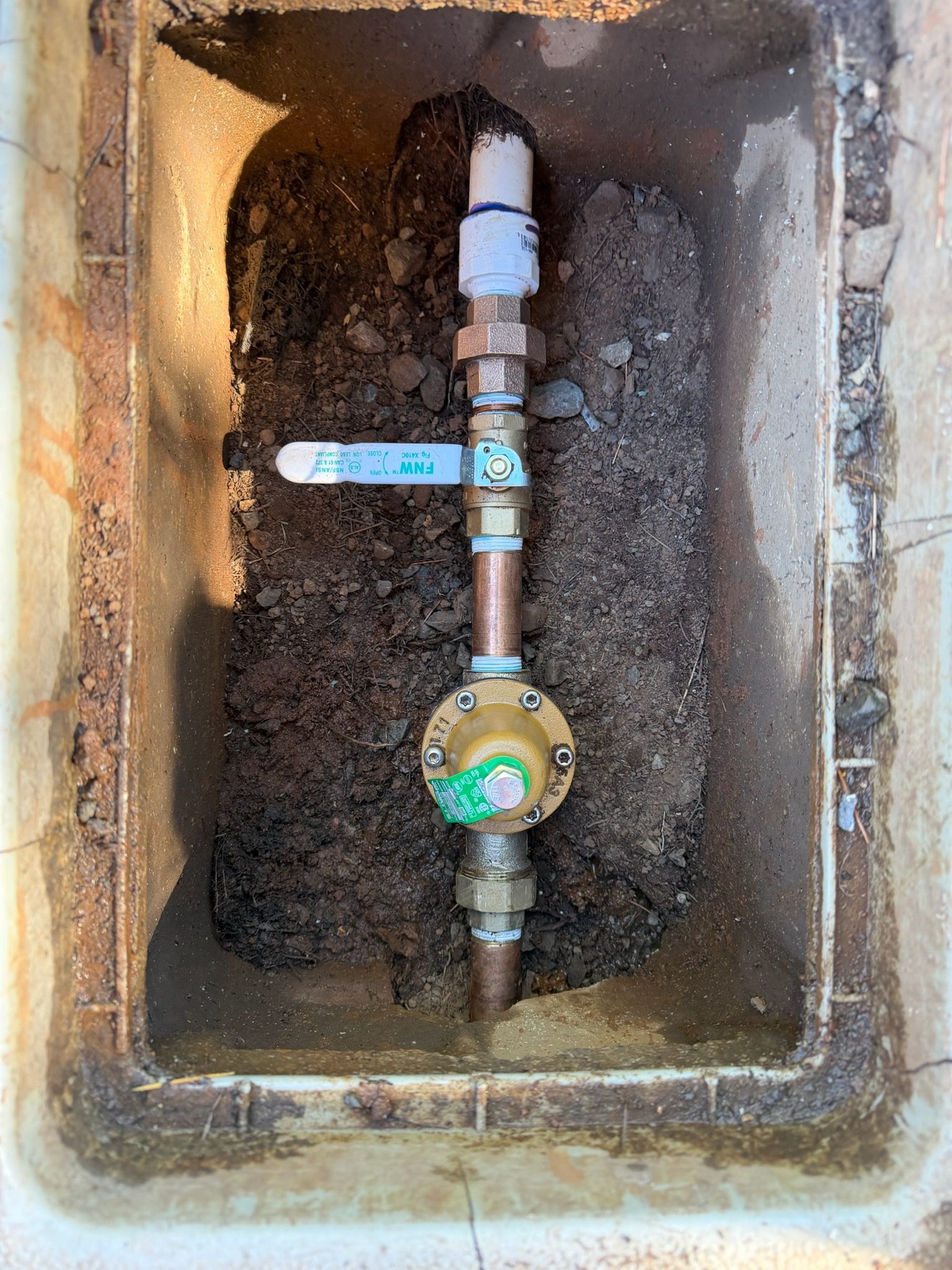 Open utility box with water meter, pipes, and shutoff valve in a dirt-lined pit