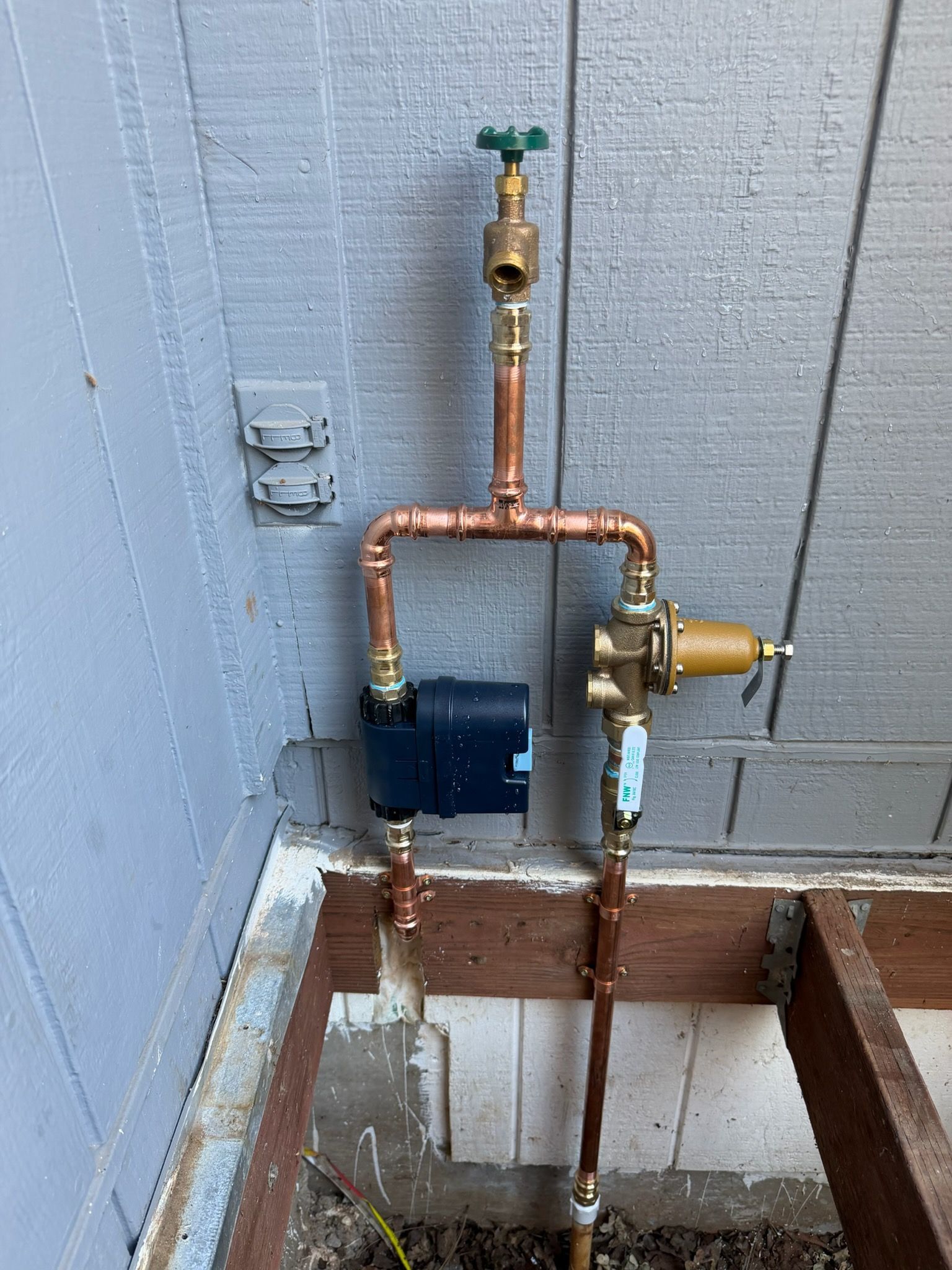 Copper plumbing pipes with valves and a blue water meter on a wall exterior.