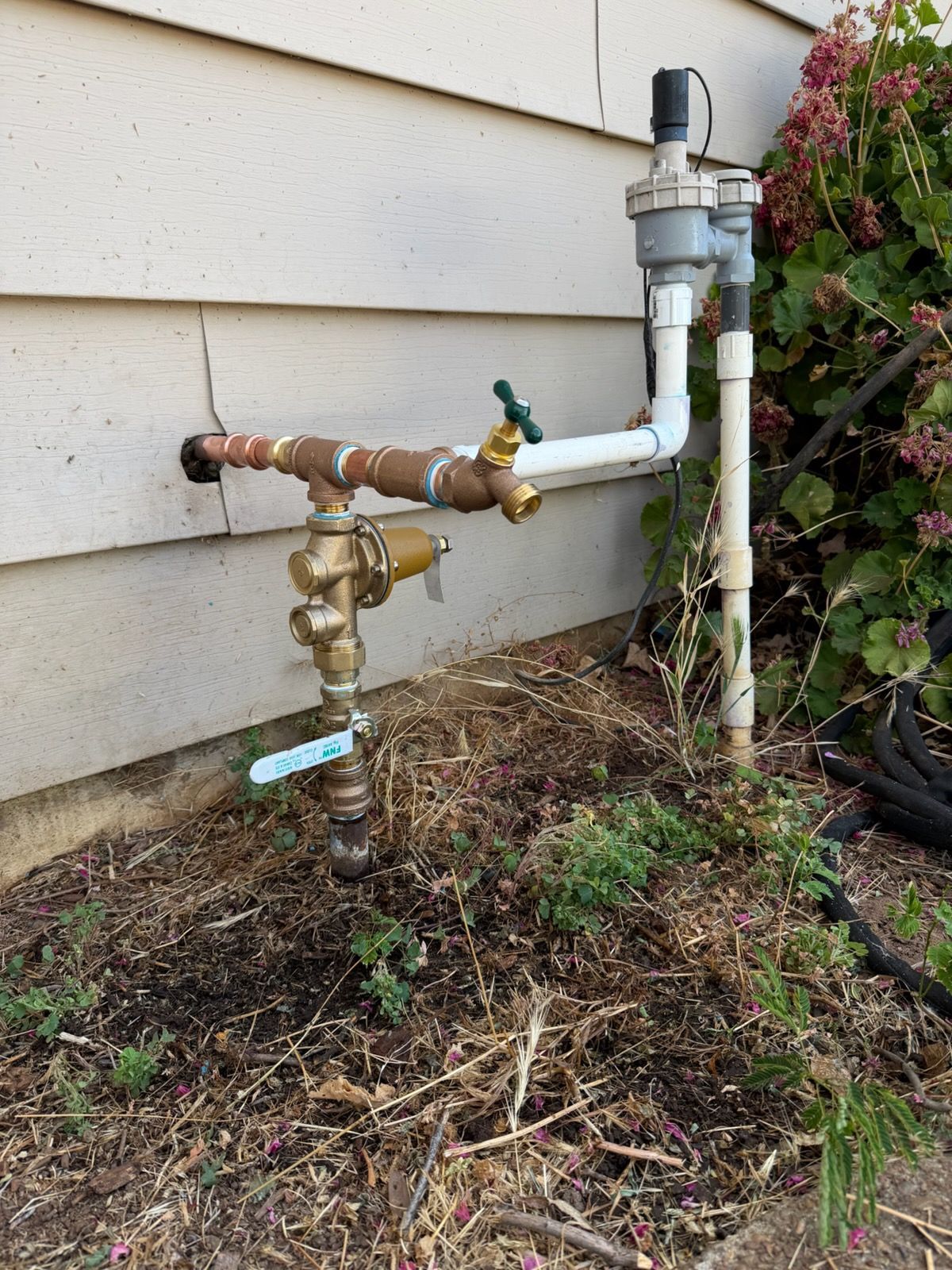 Outdoor water meter and shutoff valve with white piping beside a house foundation and shrubbery