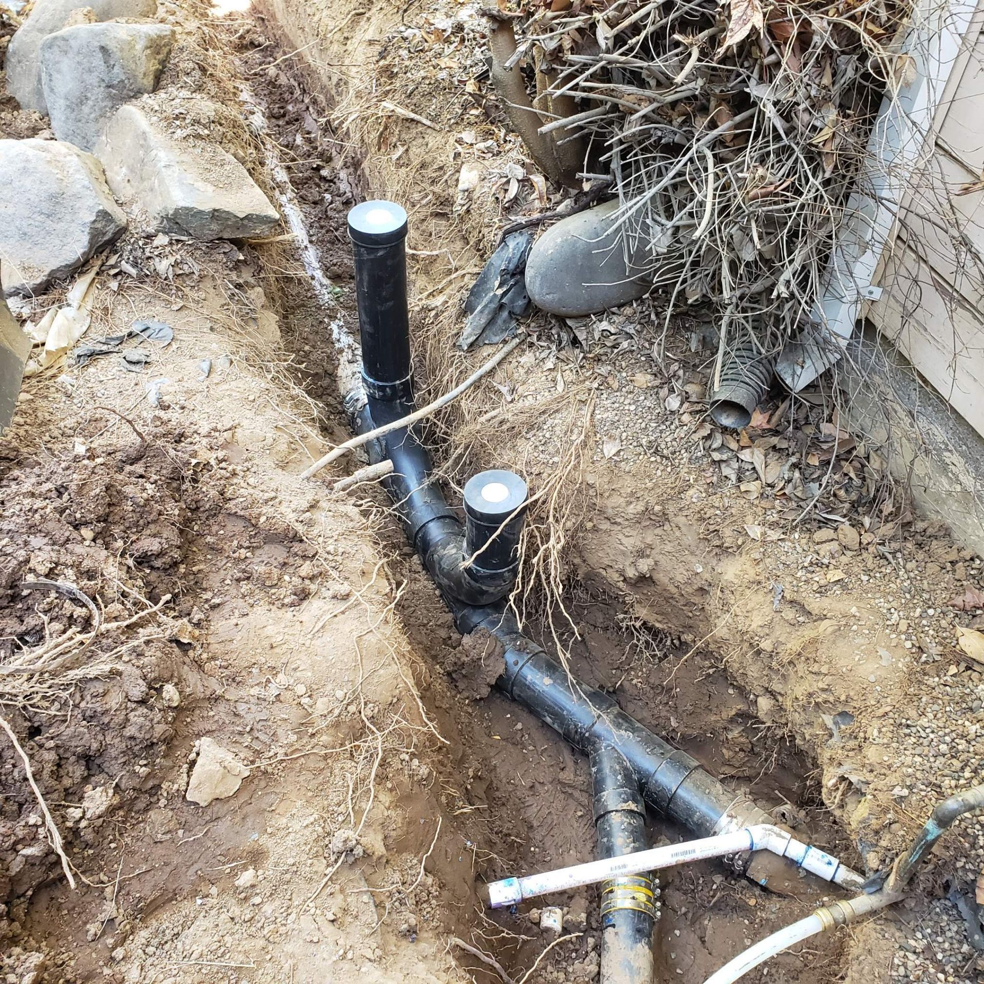 Black pipes in a trench, likely for drainage, near a building foundation and debris.
