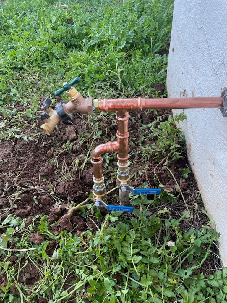 Copper water pipes and faucets installed near a concrete wall, in a grassy, dirt area.