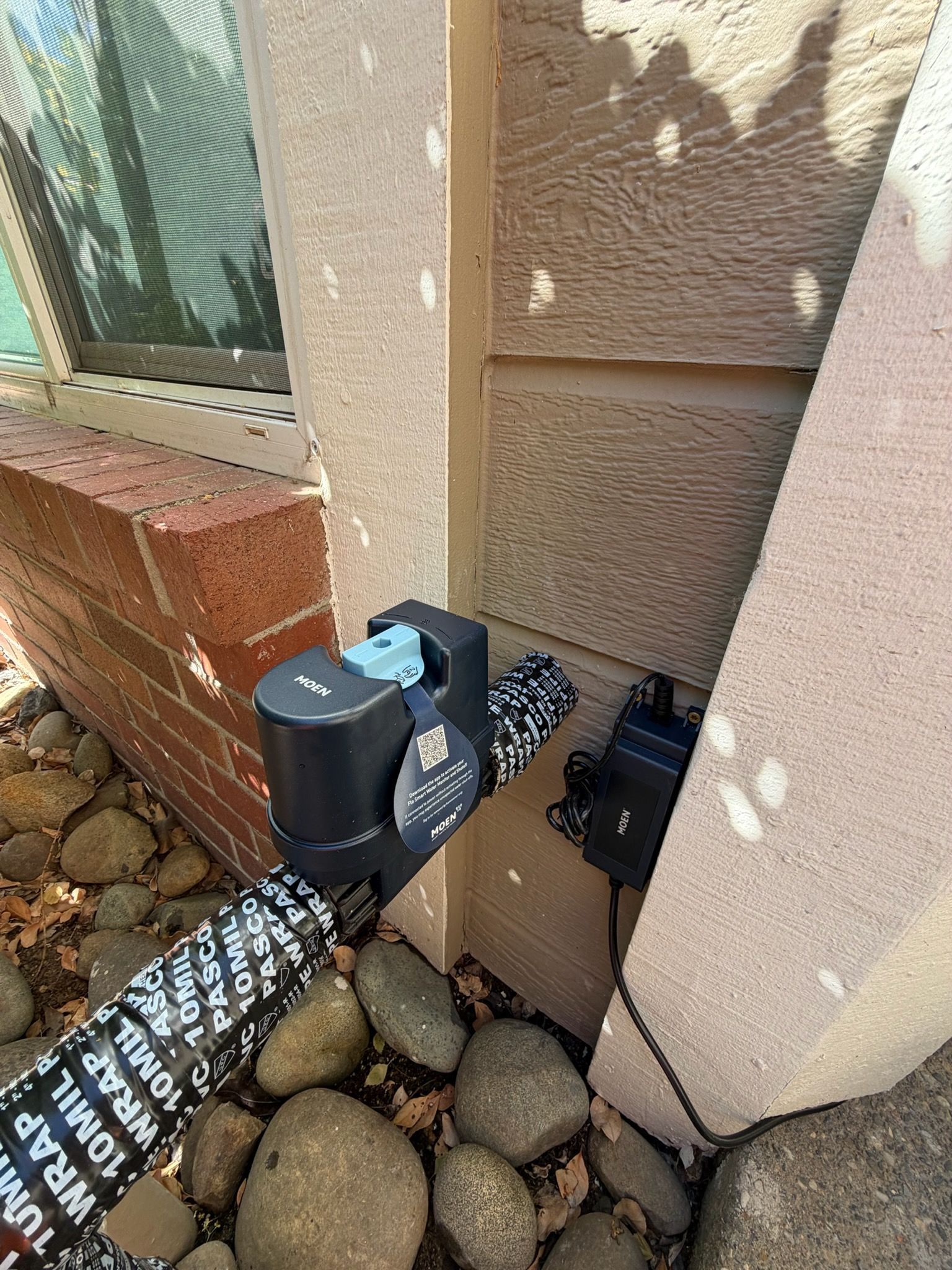 Black outdoor valve or sensor mounted beside a stucco wall over decorative rocks.