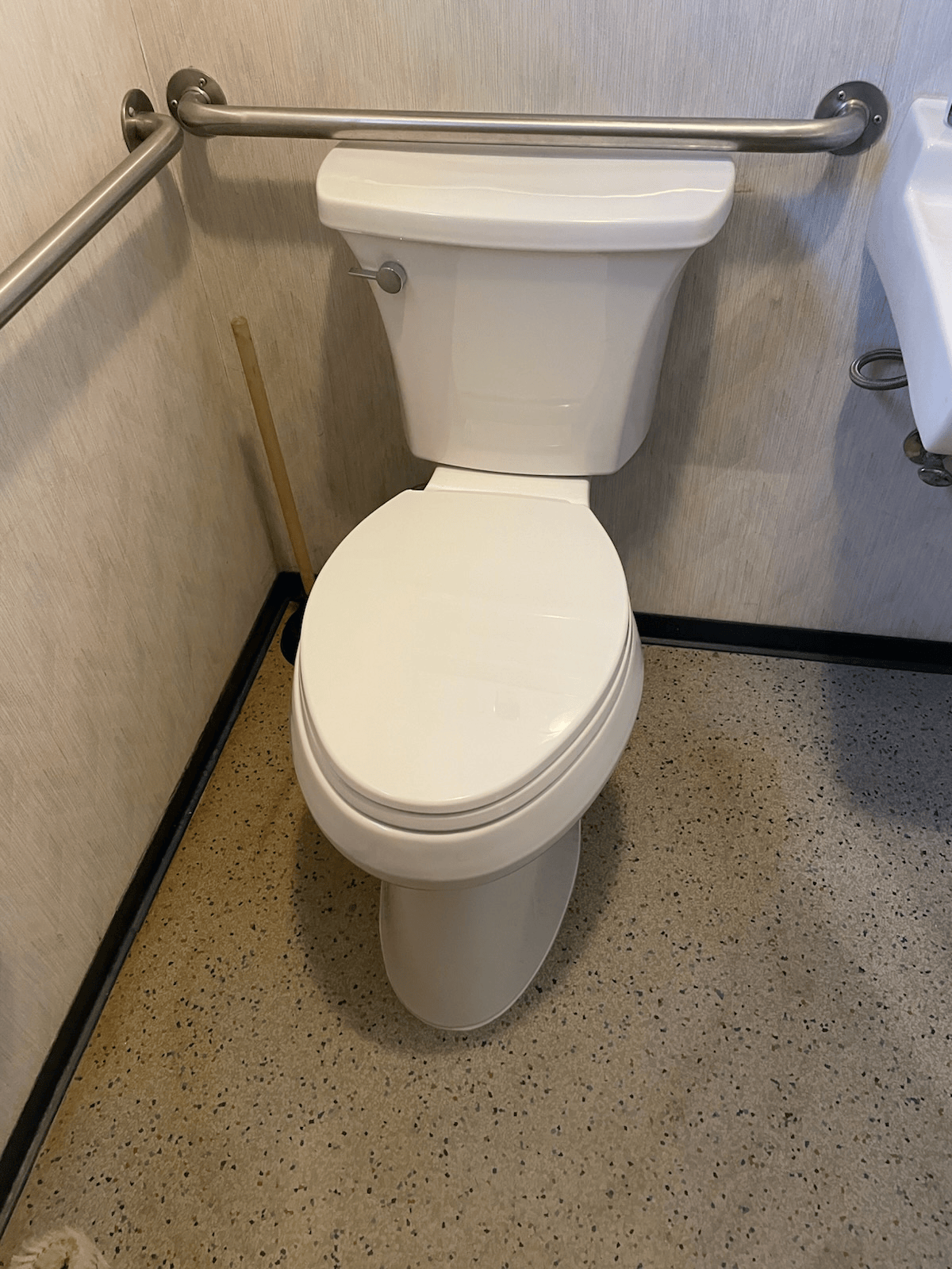 White toilet in a public restroom with a silver grab bar and speckled gray floor.
