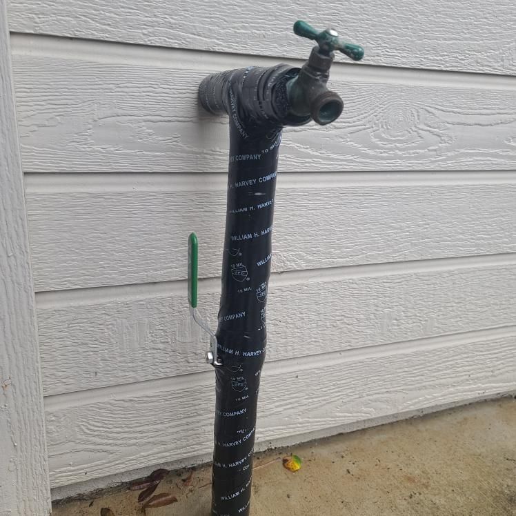 Outdoor water faucet, black-taped pipe, mounted on a white siding wall.