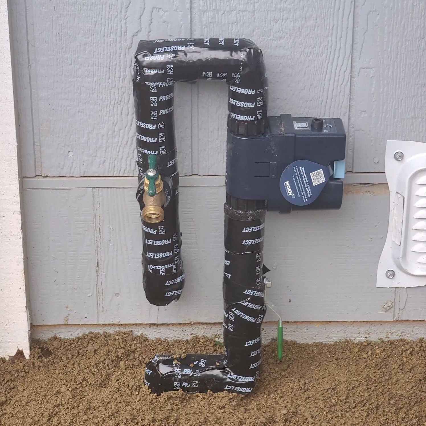 Wrapped outdoor pipes and a water meter against a house wall, insulated with black foam.