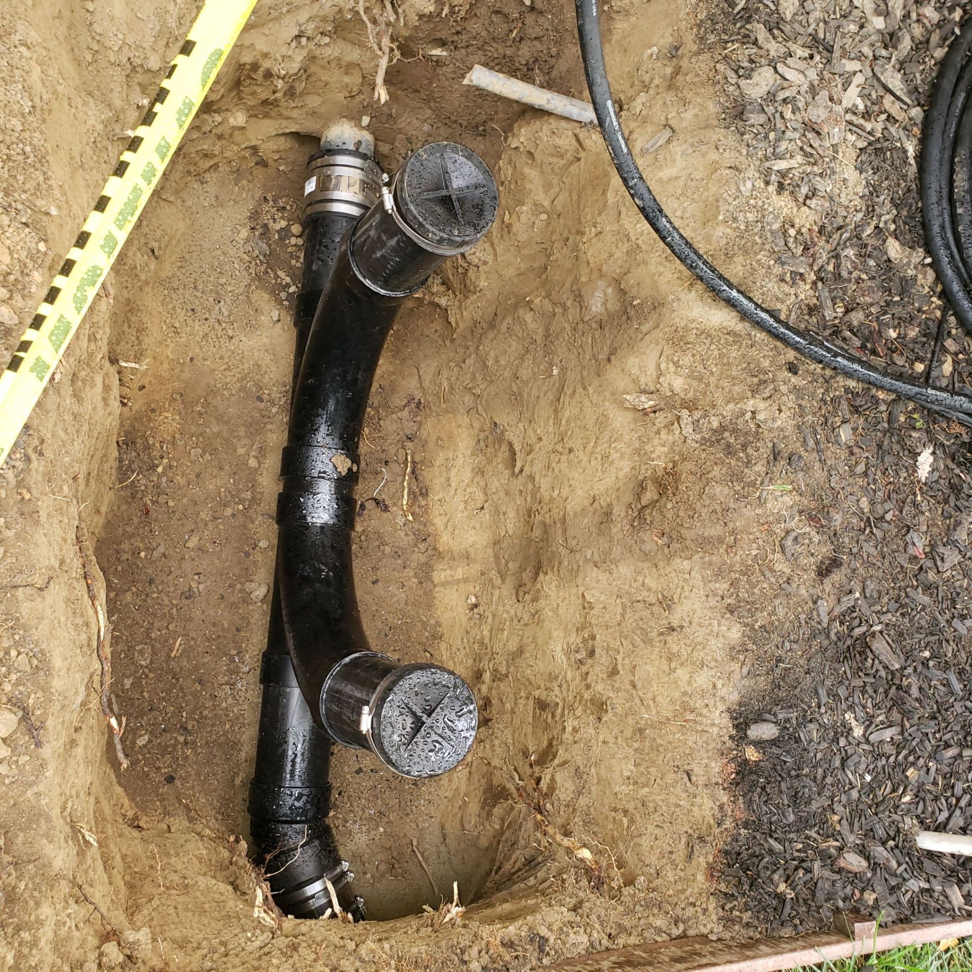 Black plumbing pipes in a dirt hole. Two capped pipes at the top. Yellow measuring tape.