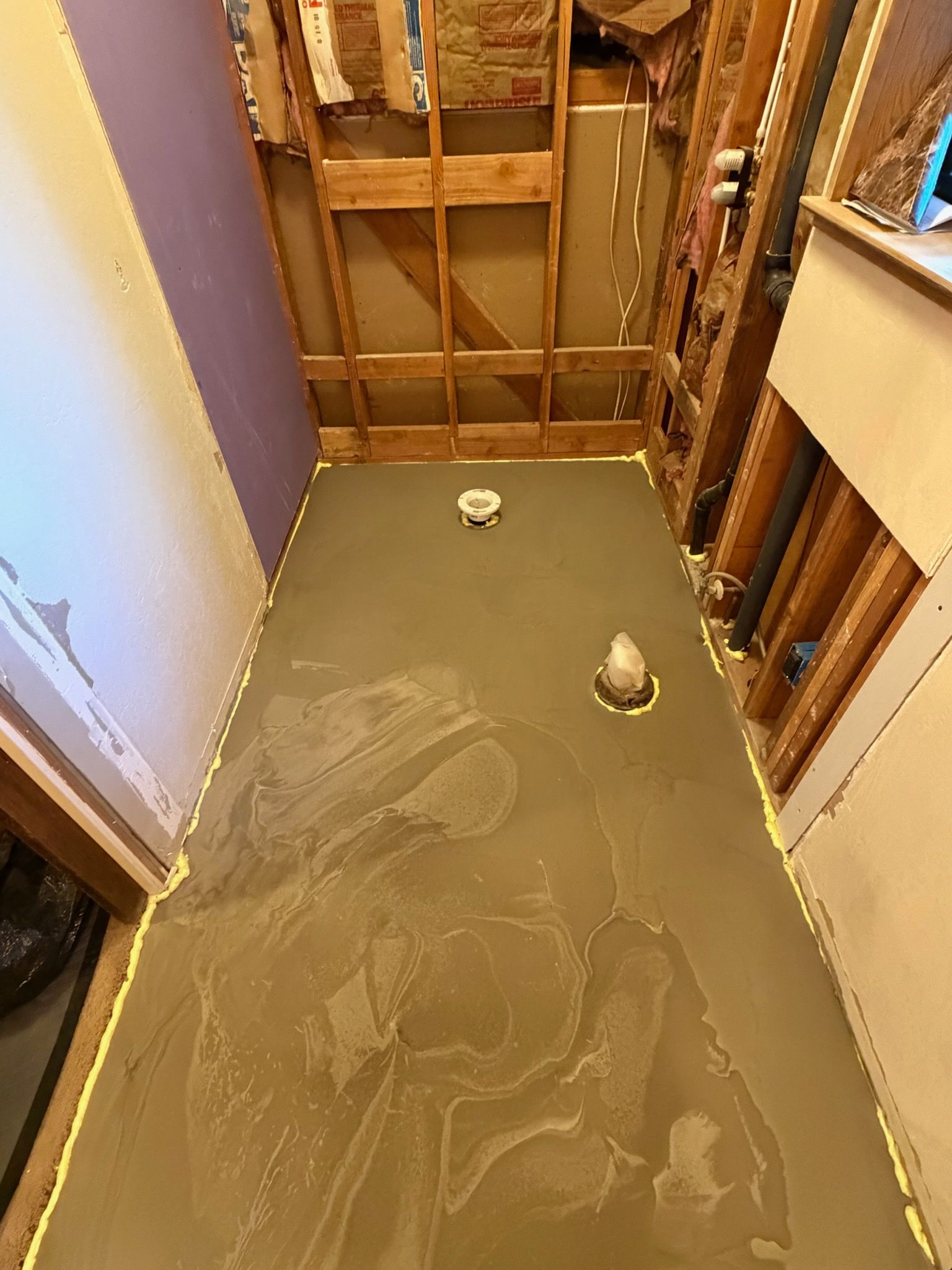 Partially demolished bathroom floor with exposed wall studs and plumbing, dusty concrete surface