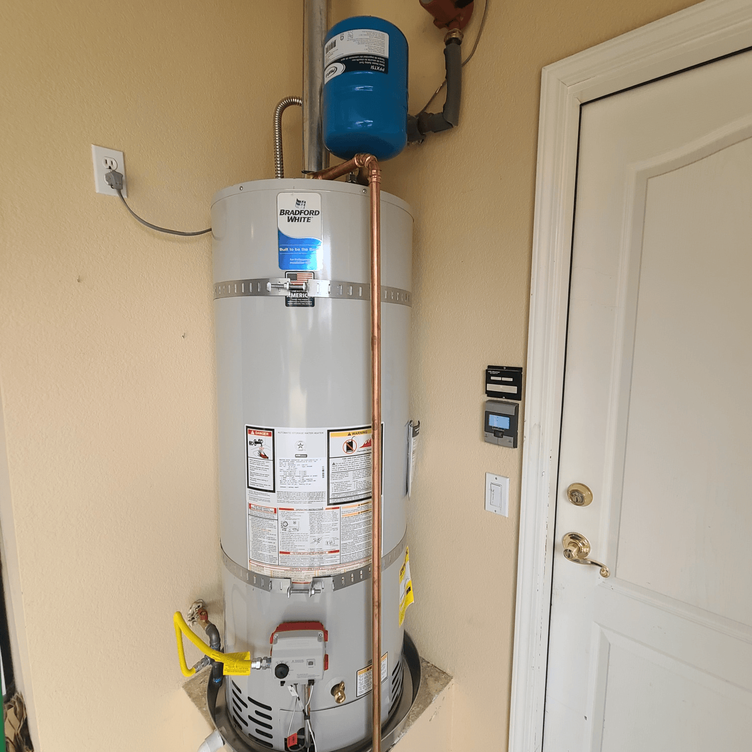 Water heater against beige wall, with a blue expansion tank on top and a white door to the right.