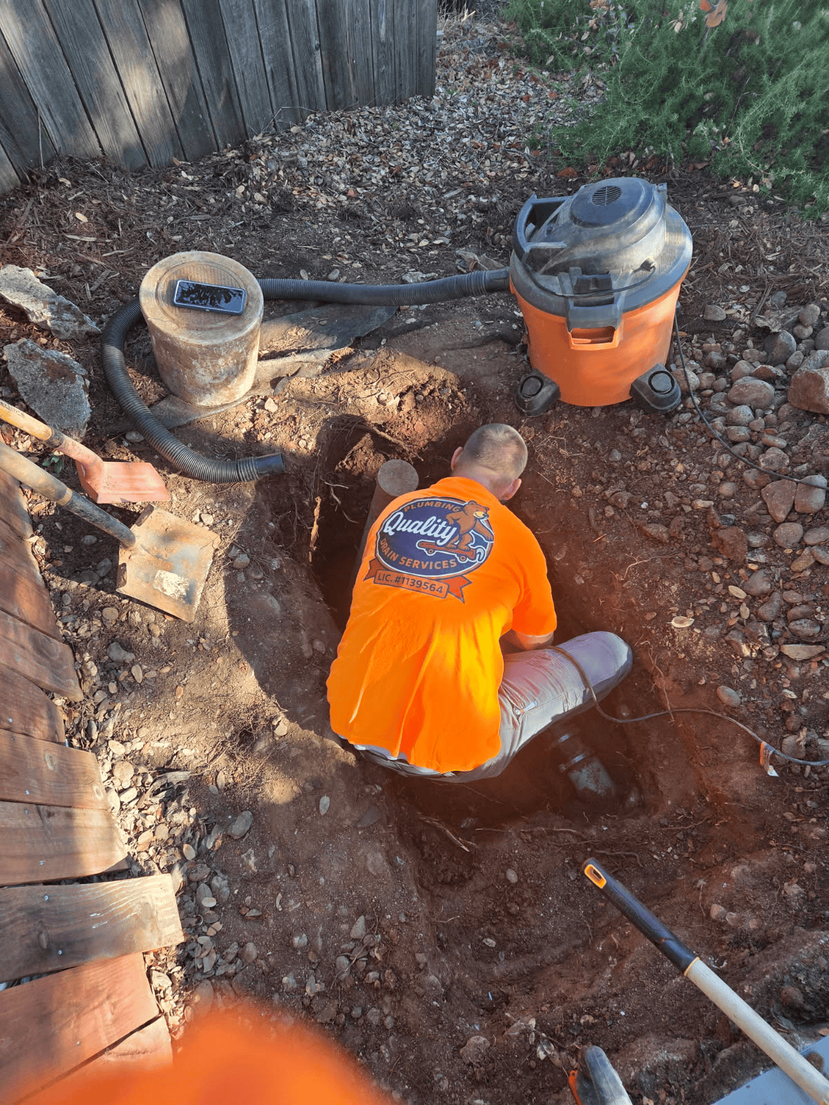 A person wearing an orange shirt is in a deep hole, possibly repairing something outdoors, with a vacuum nearby.