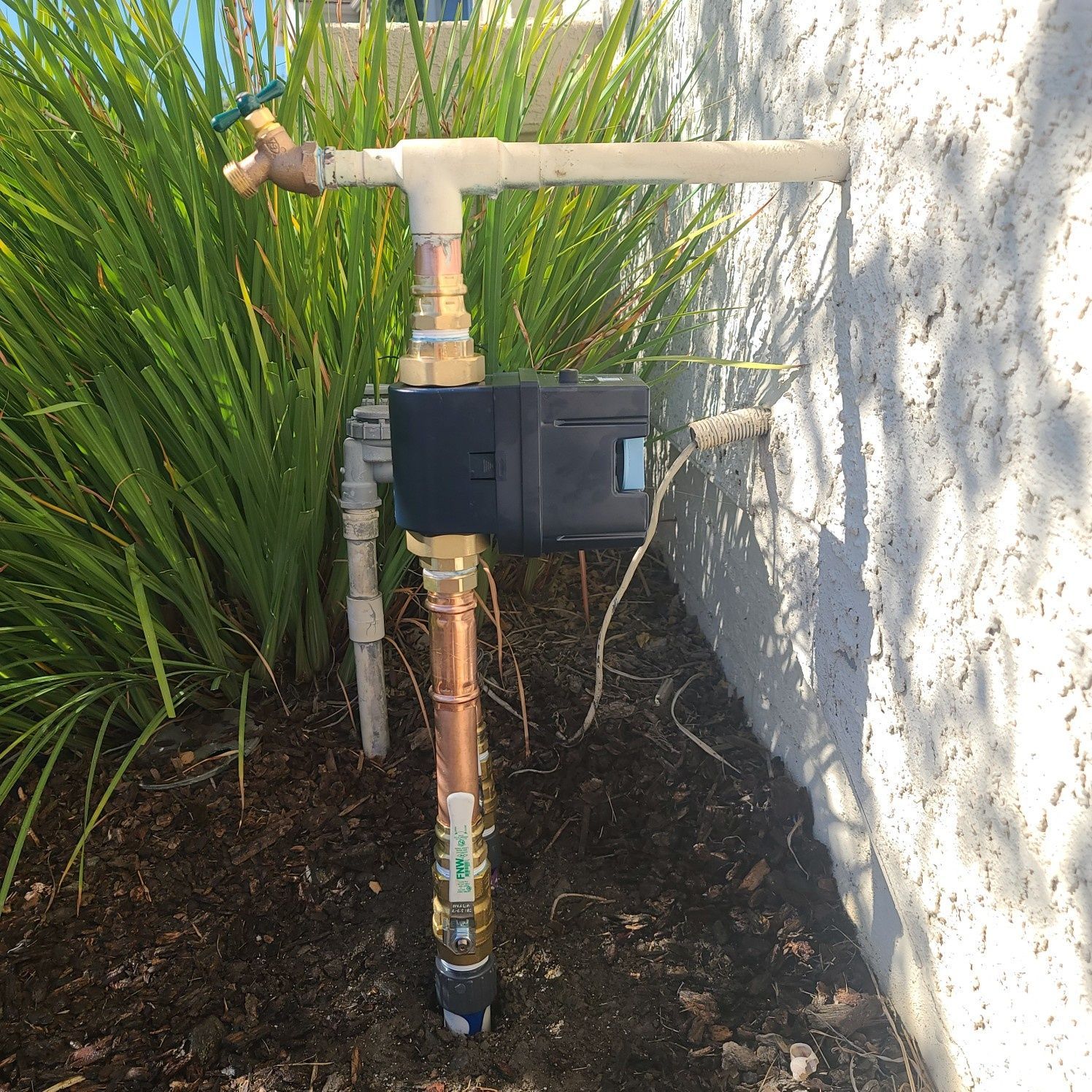 Outdoor water meter with pipes and spigot against a stucco wall, with green plants.