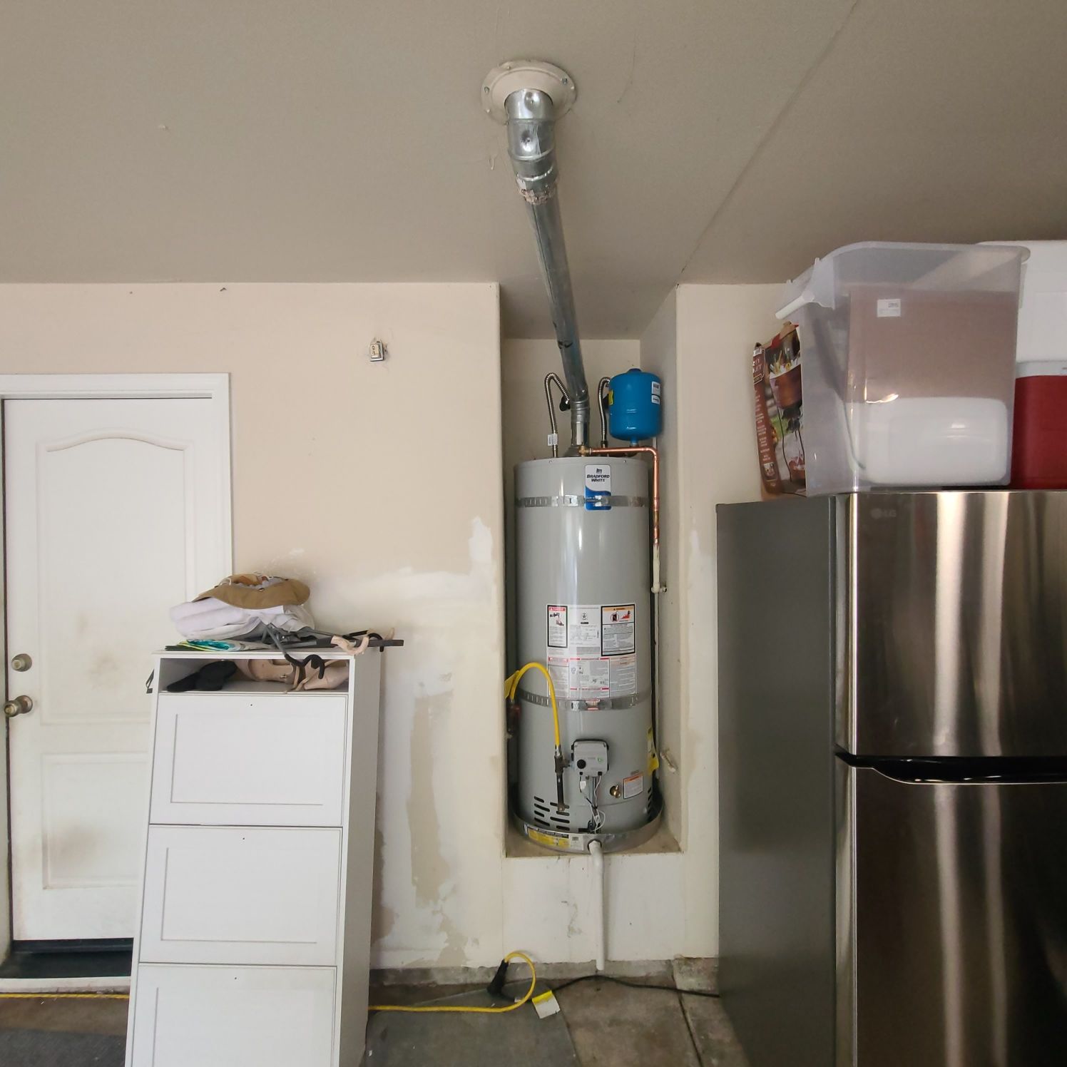 A water heater in a garage alcove with a long, angled pipe leading to the ceiling. A blue expansion tank is on the side.