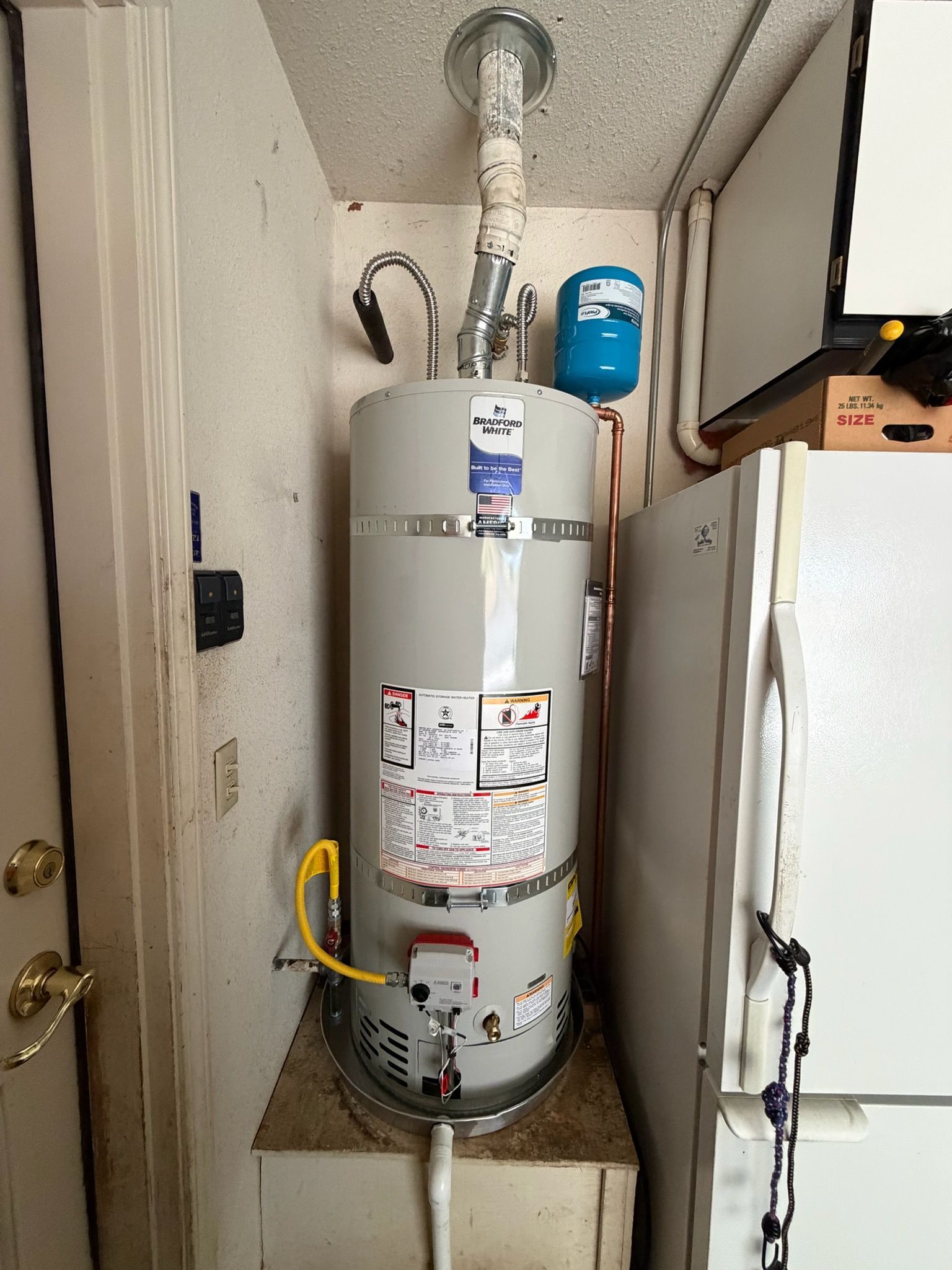 Water heater in a narrow utility closet with exposed piping and a nearby blue expansion tank.