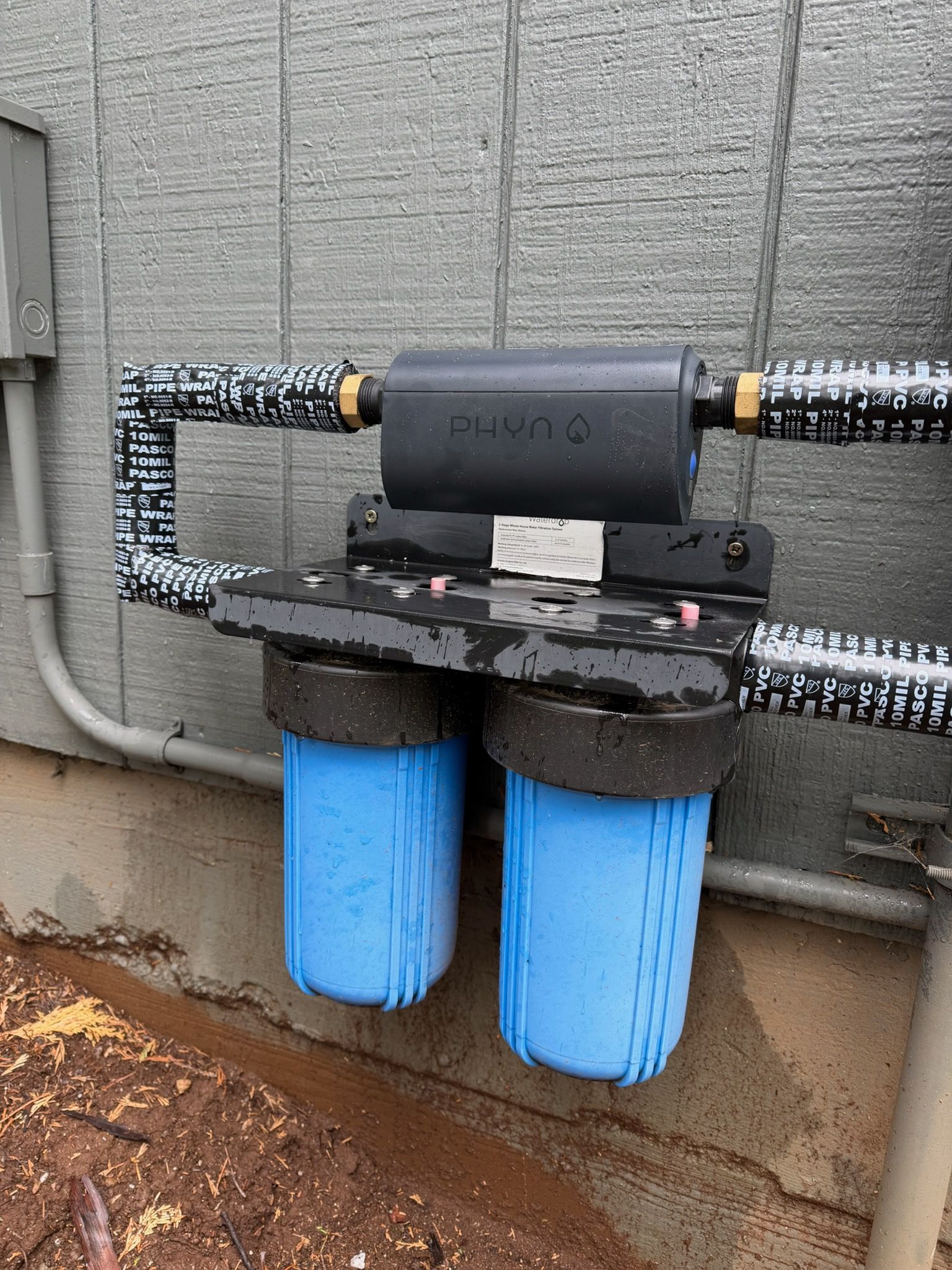 Two blue inline water filters mounted on a wall, connected to black pipes.