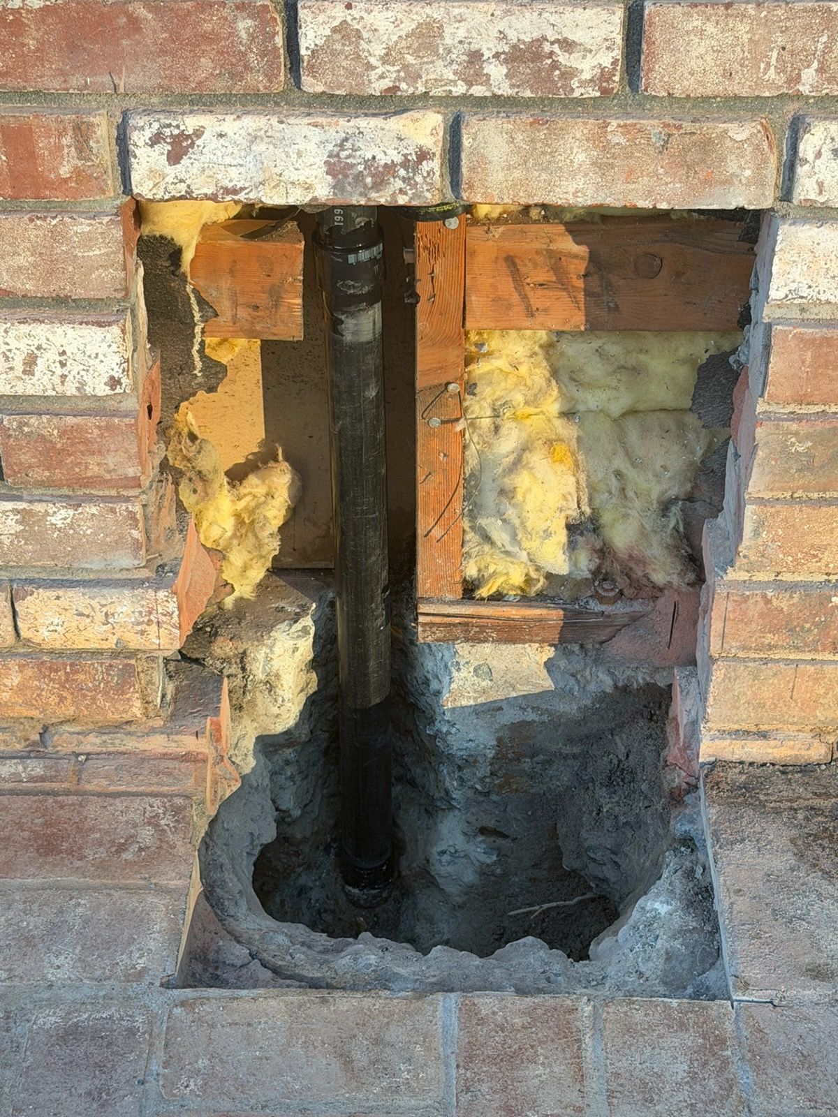 Hole cut into a brick wall, revealing a pipe, insulation, and brick remnants.