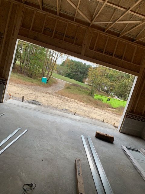 Construction site: open garage door frame looking out onto a dirt path, trees, and grass; unfinished concrete floor.