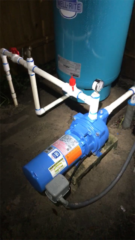 A blue well pump connected to white PVC pipes and a blue Well-Rite pressure tank on a dirt floor.