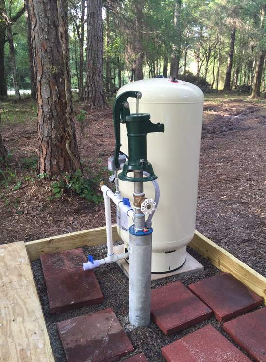 A water well pump and beige pressure tank stand on a wooden-framed gravel pad in a wooded outdoor setting.
