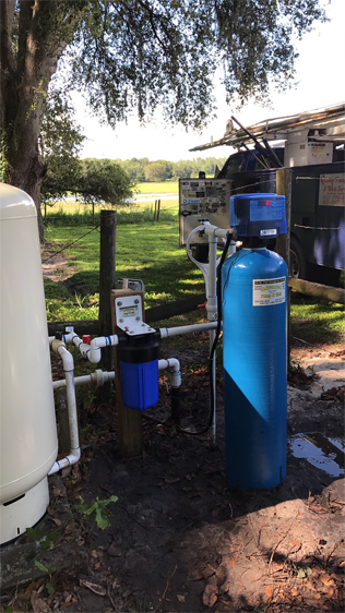 Outdoor well water filtration system featuring a tall white pressure tank, a blue filter housing, and a blue water softener.