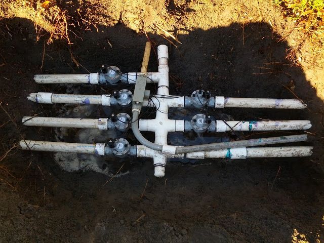 A PVC pipe sprinkler manifold with multiple valves and wiring buried in a dirt trench.