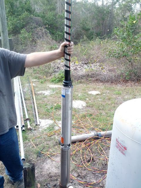A person holding a stainless steel submersible water pump attached to a pipe in an outdoor setting.