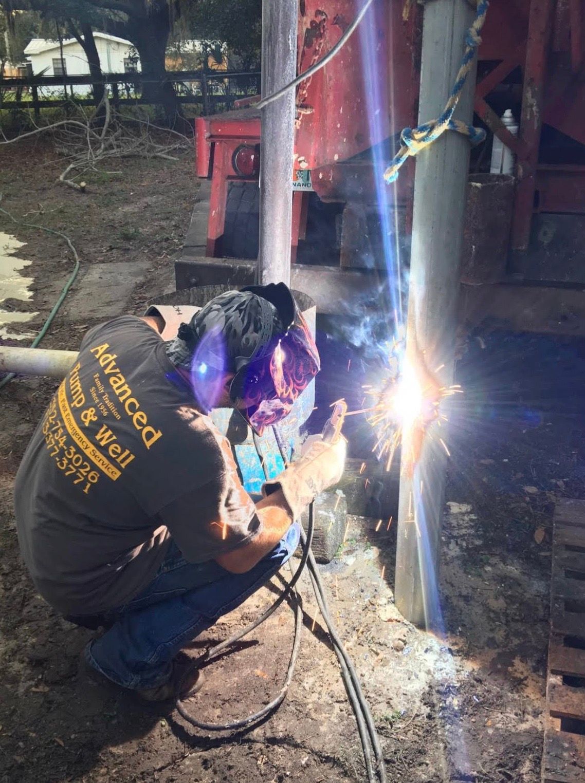 A welder in a dark shirt performs repairs on a metal pipe outdoors, with sparks visible from the welding arc.